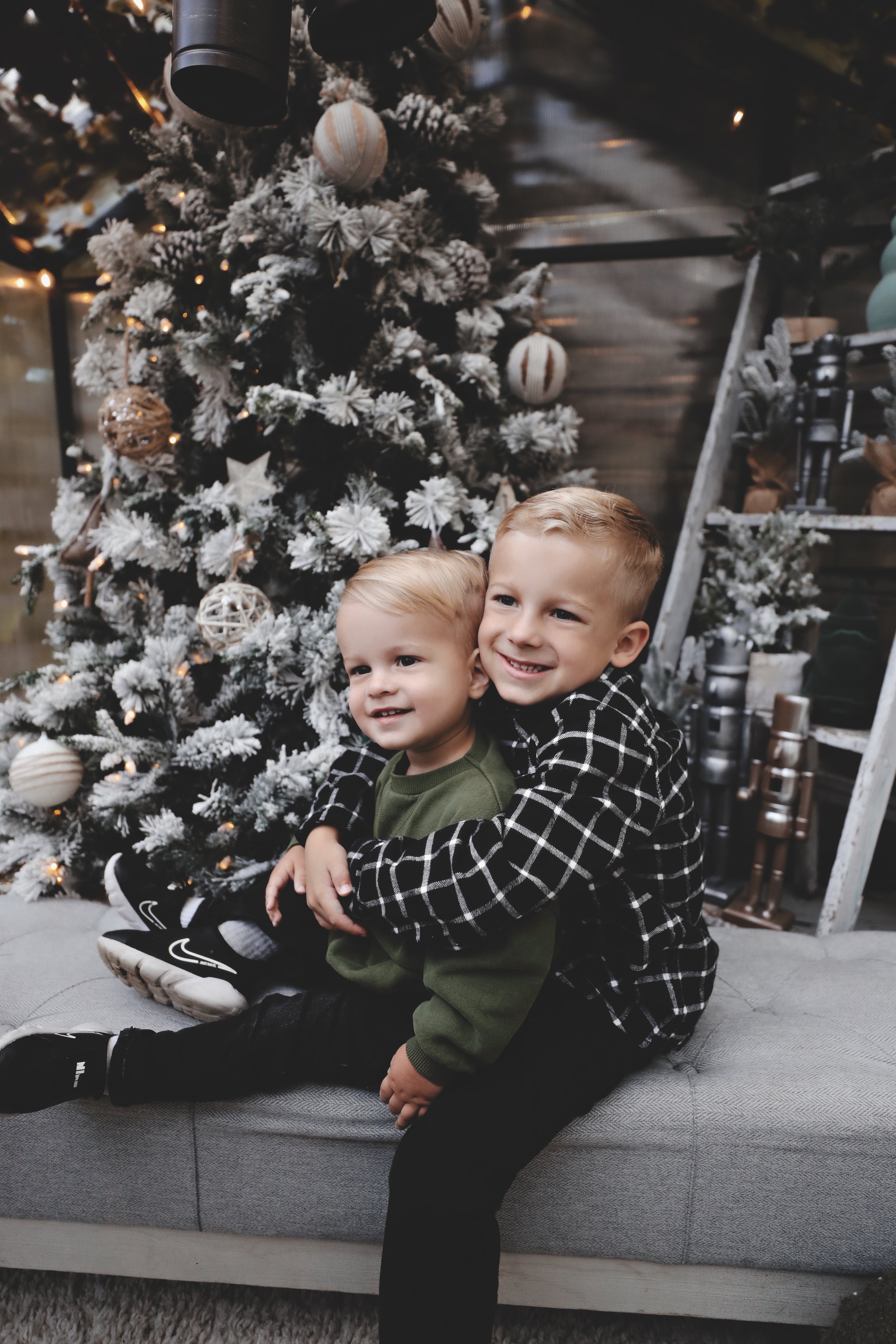 Two young boys sitting on a gray bench hugging in front of a decorated Christmas tree with white and beige ornaments, in a cozy indoor setting.