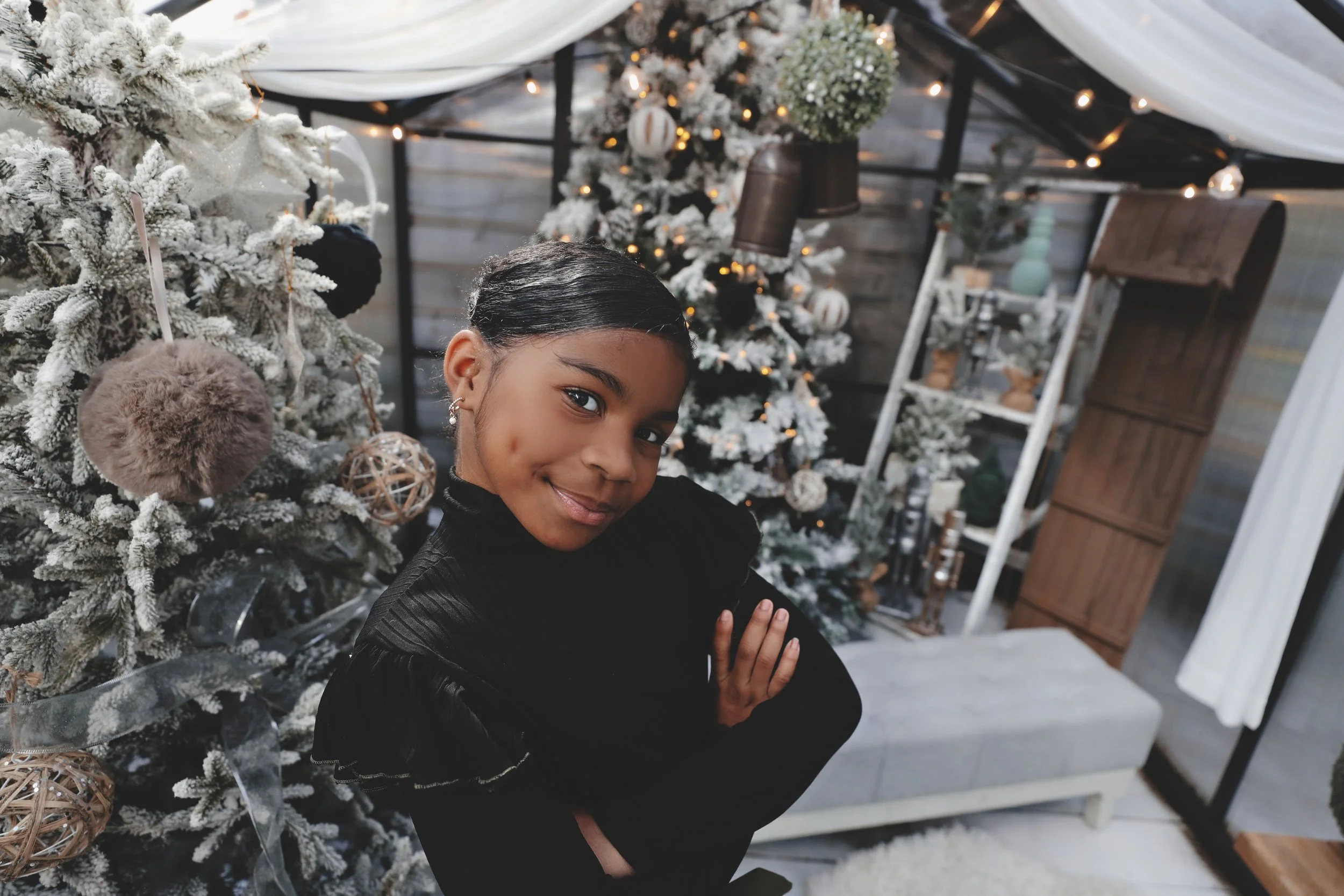 A young girl with black hair and a black top smiling with her arms crossed, posing near a decorated Christmas tree in a cozy room.