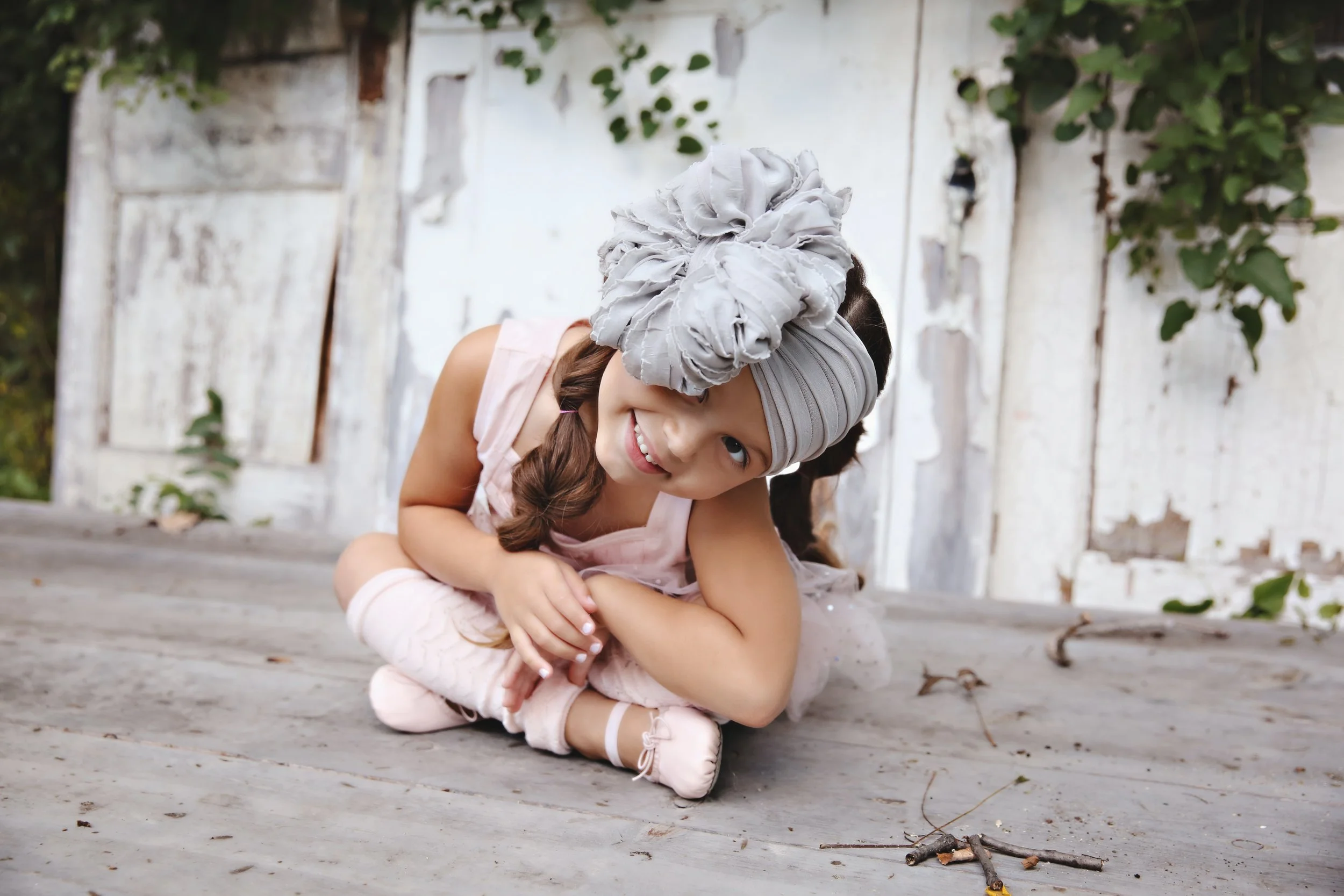 A young girl in ballet outfit sitting on a wooden floor, smiling, with white open-toed ballet slippers. She is wearing a grey headwrap with a large fabric flower on top, a pink sleeveless dress, with a rustic white wooden background and some green fo