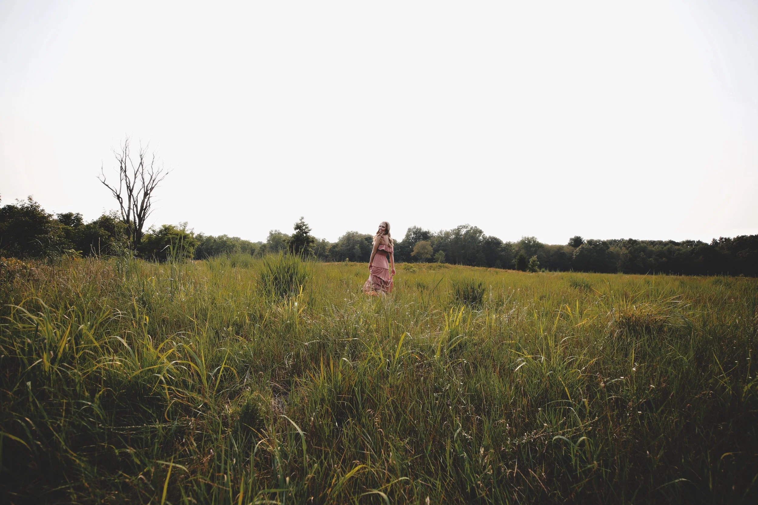 A woman in a pink dress standing in a grassy field with trees in the background under a bright sky.
