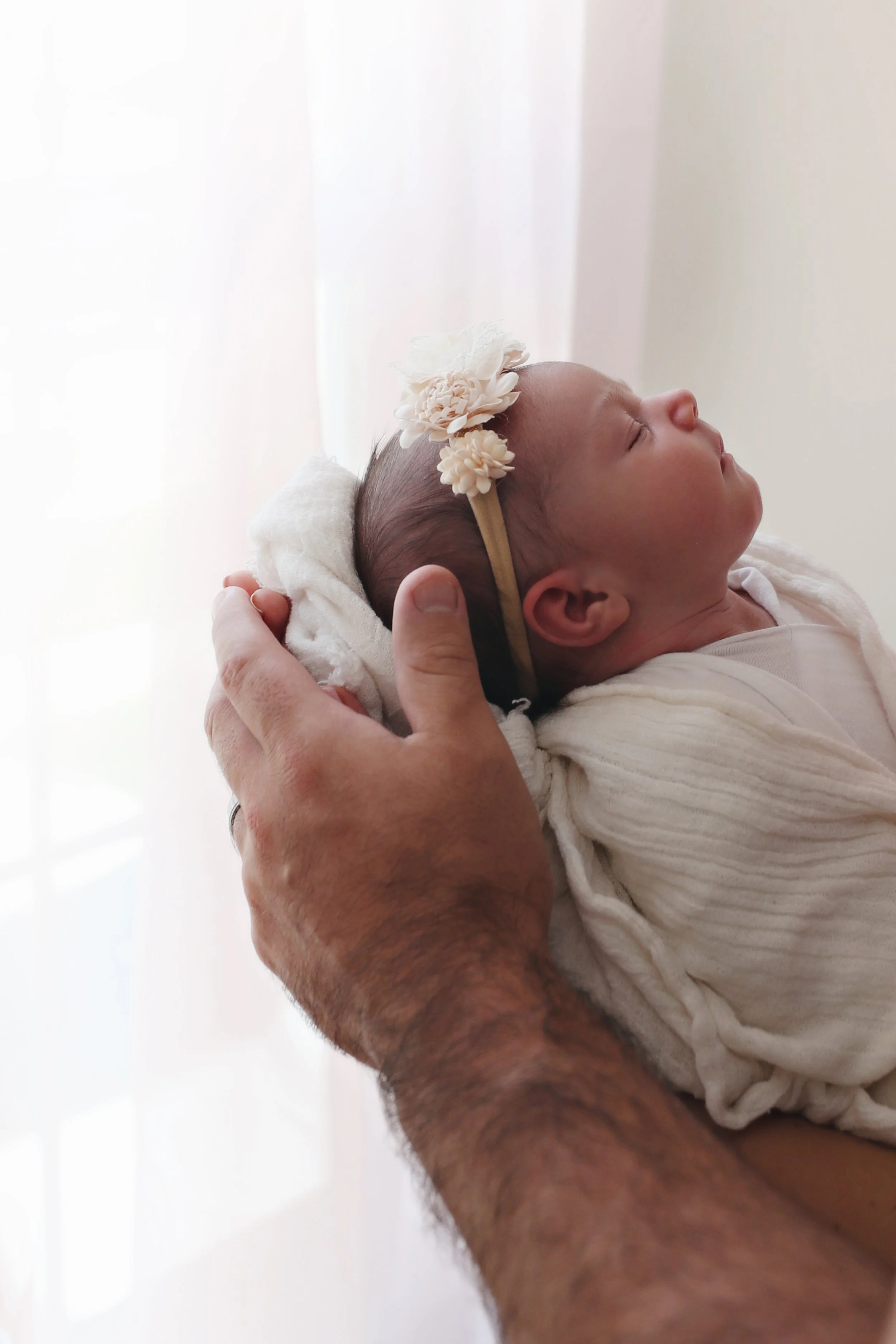 Close-up of a newborn baby girl with a floral headband, sleeping peacefully in an adult's arms against a bright background.