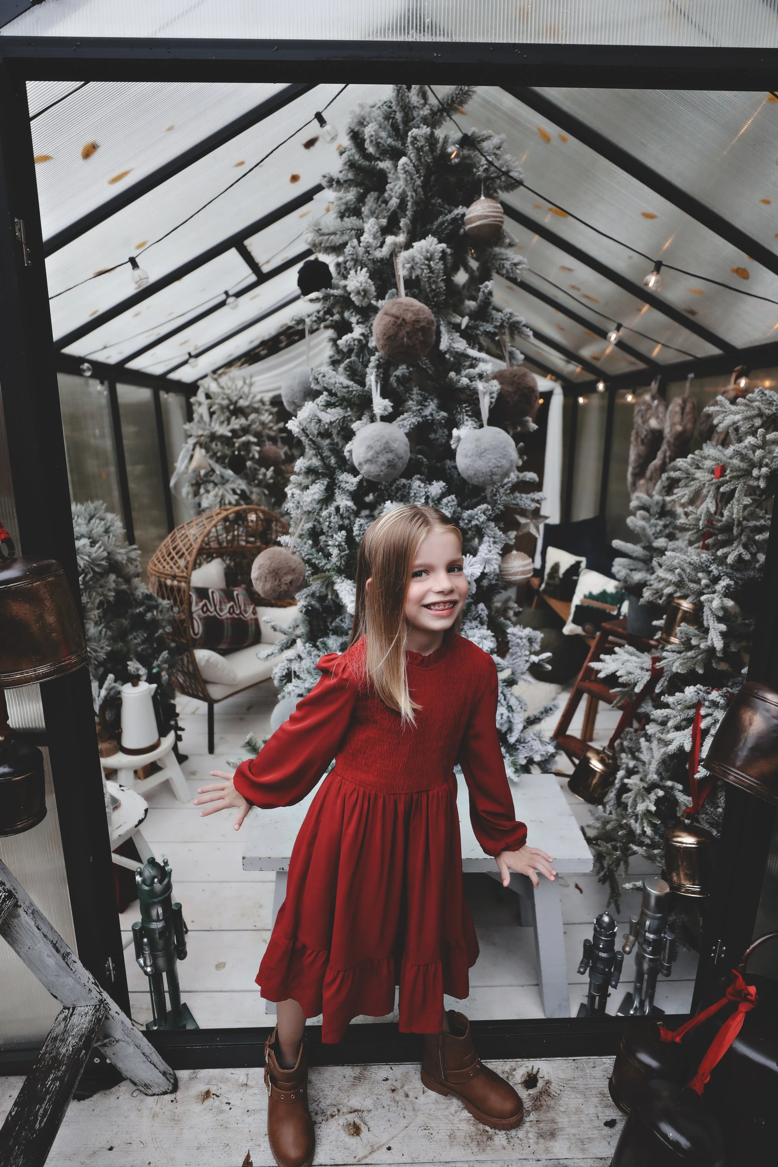 A young girl in a red dress and brown boots standing in front of a snow-flocked Christmas tree inside a glass conservatory decorated for Christmas.