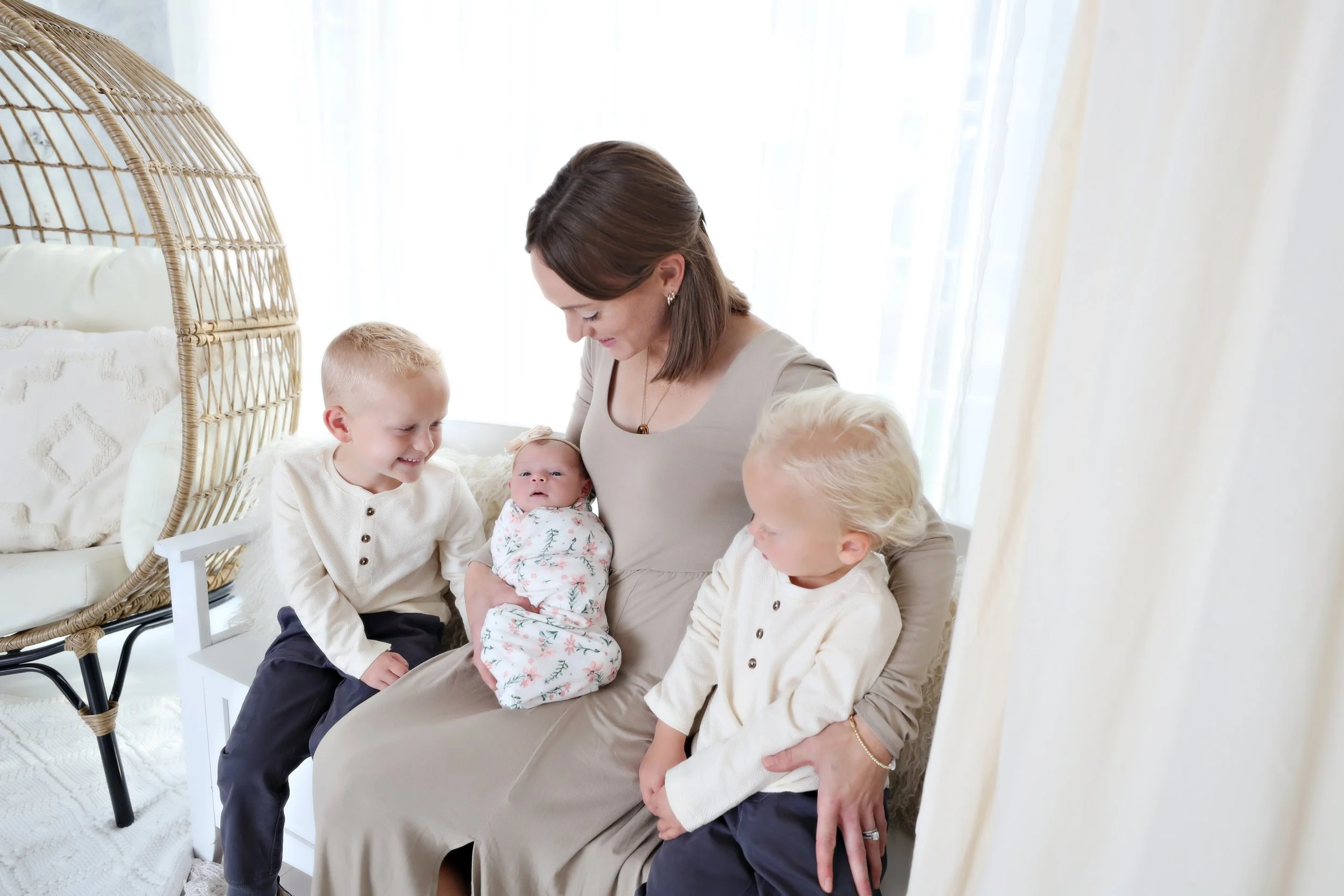 A woman sitting on a bed holding a newborn baby and smiling at two young boys, all inside a bright room with natural light.