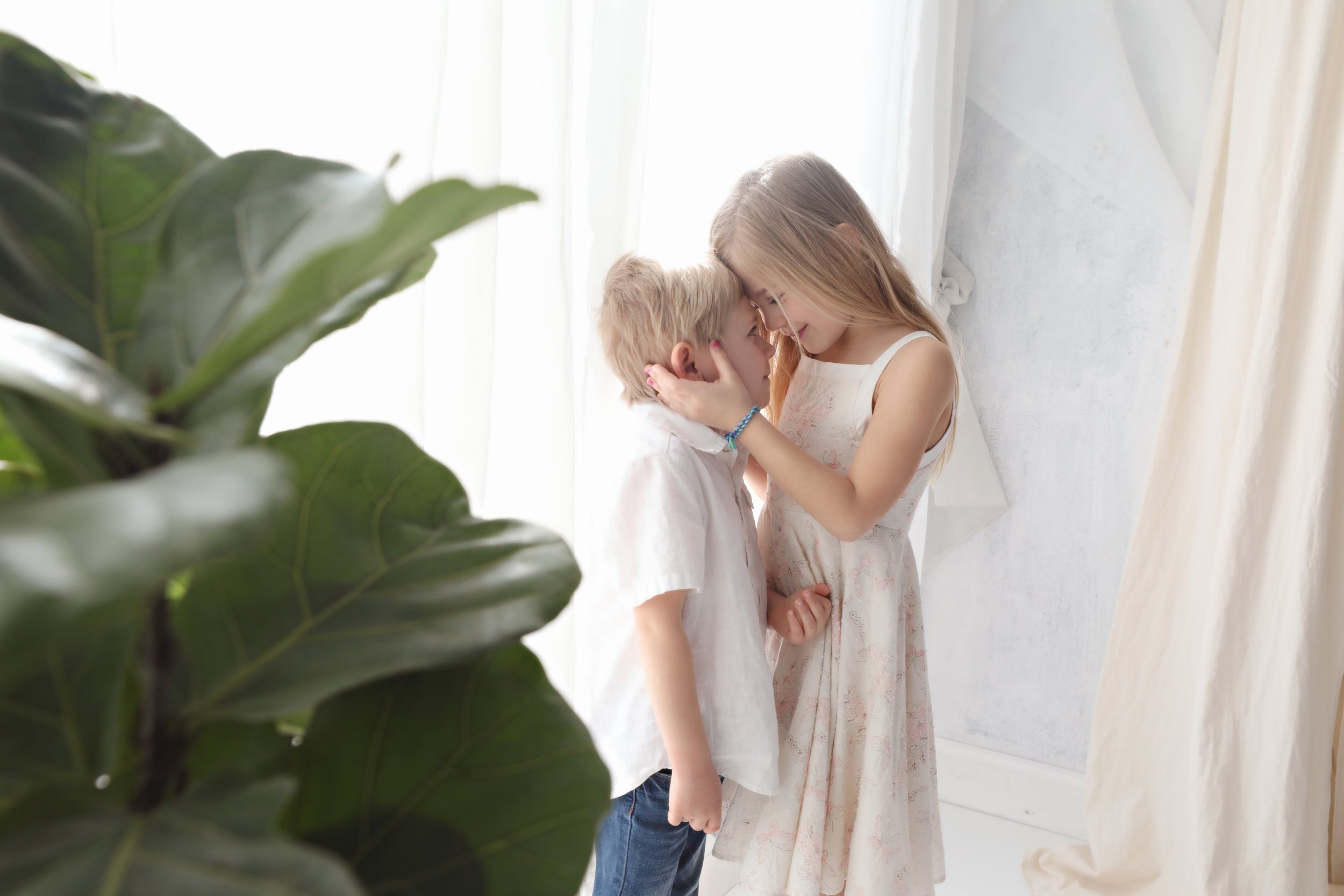A young girl and a young boy touching foreheads and smiling in a bright room, with leaves from a large plant in the foreground.