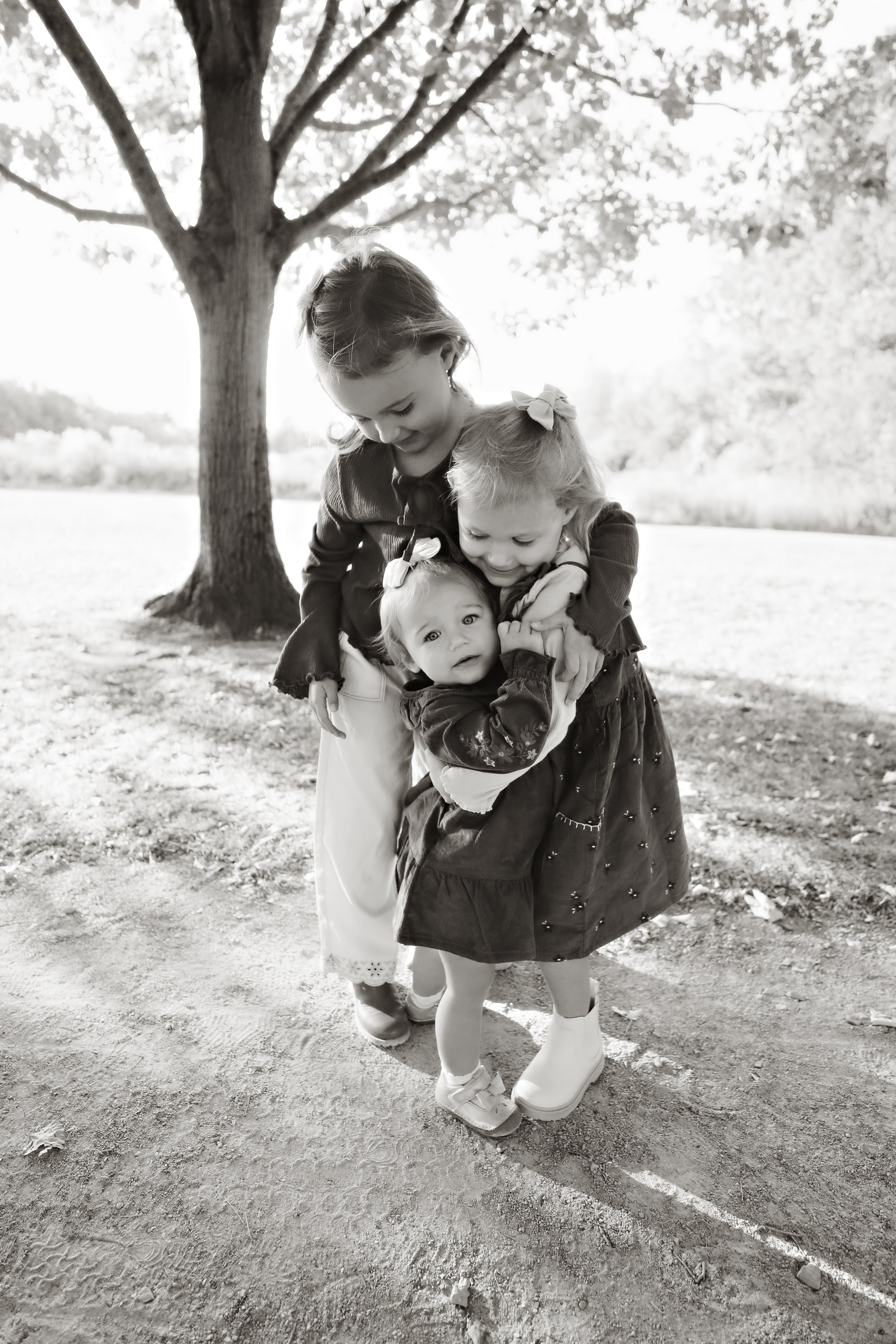 Three young girls hugging in a park near a large tree, with foliage and open space in the background.