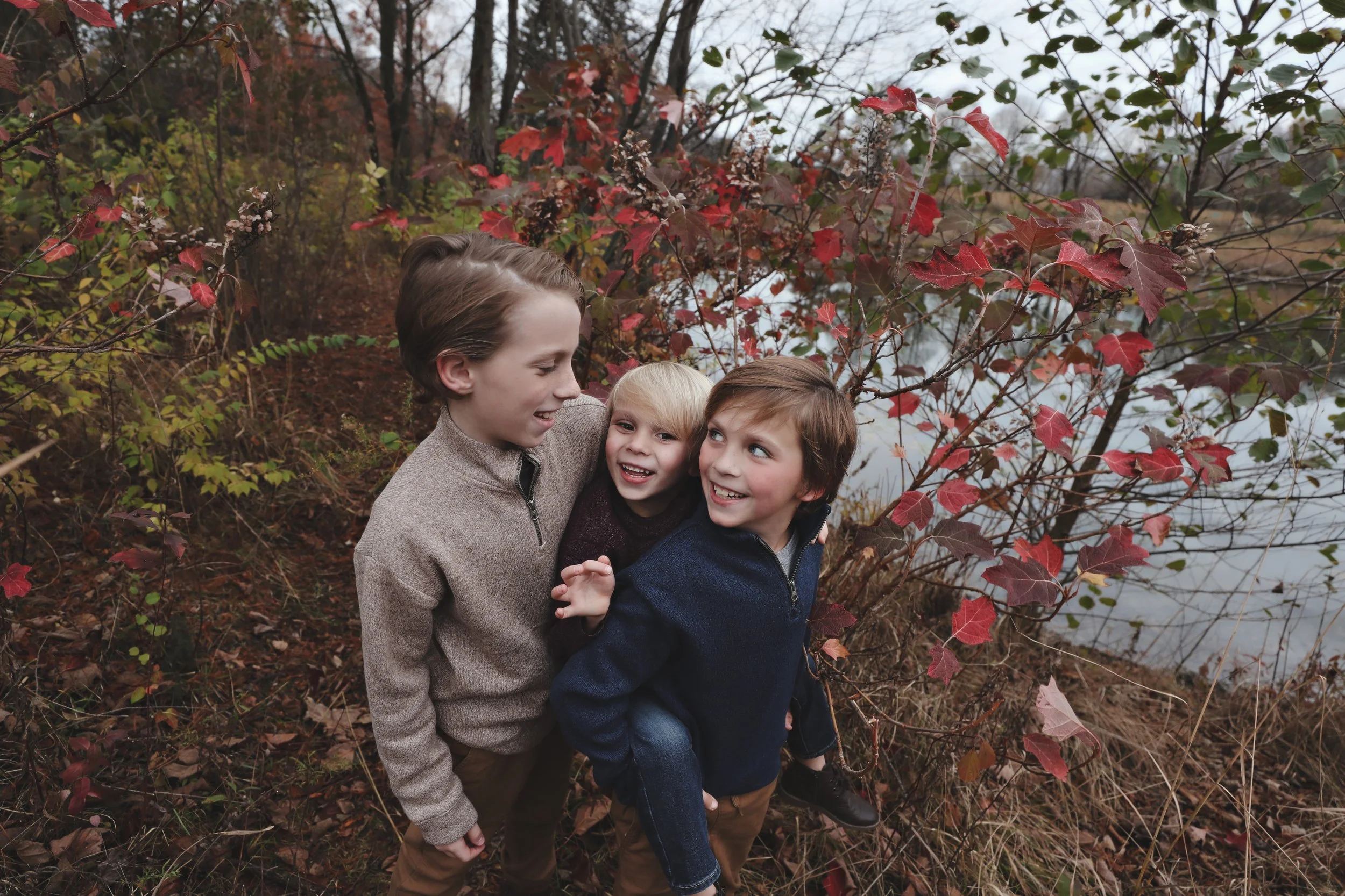 Three children playing outdoors near a lake during autumn, surrounded by trees with red and green leaves.