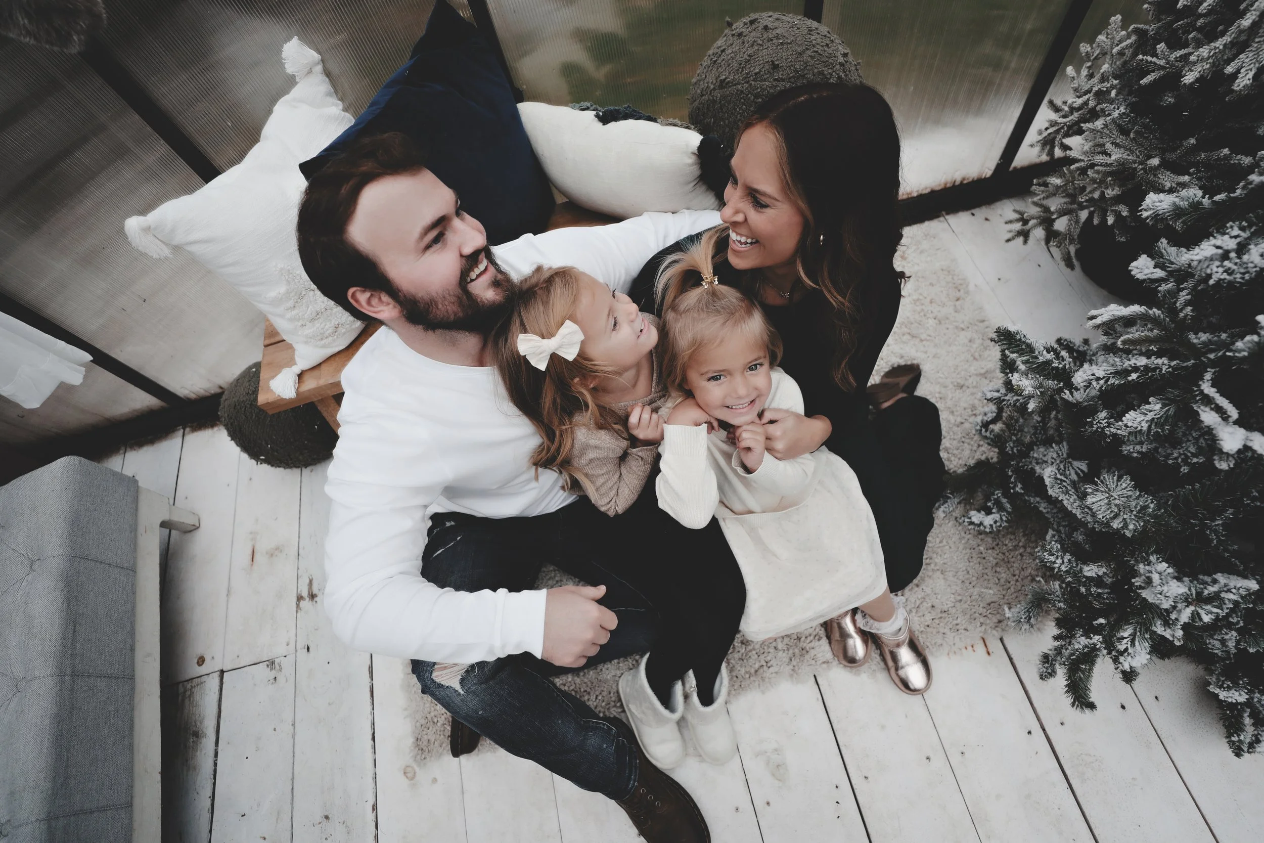 A family of four with two young girls sitting on a parent’s lap, smiling and laughing around Christmas decorations, including a Christmas tree, in a cozy indoor setting.