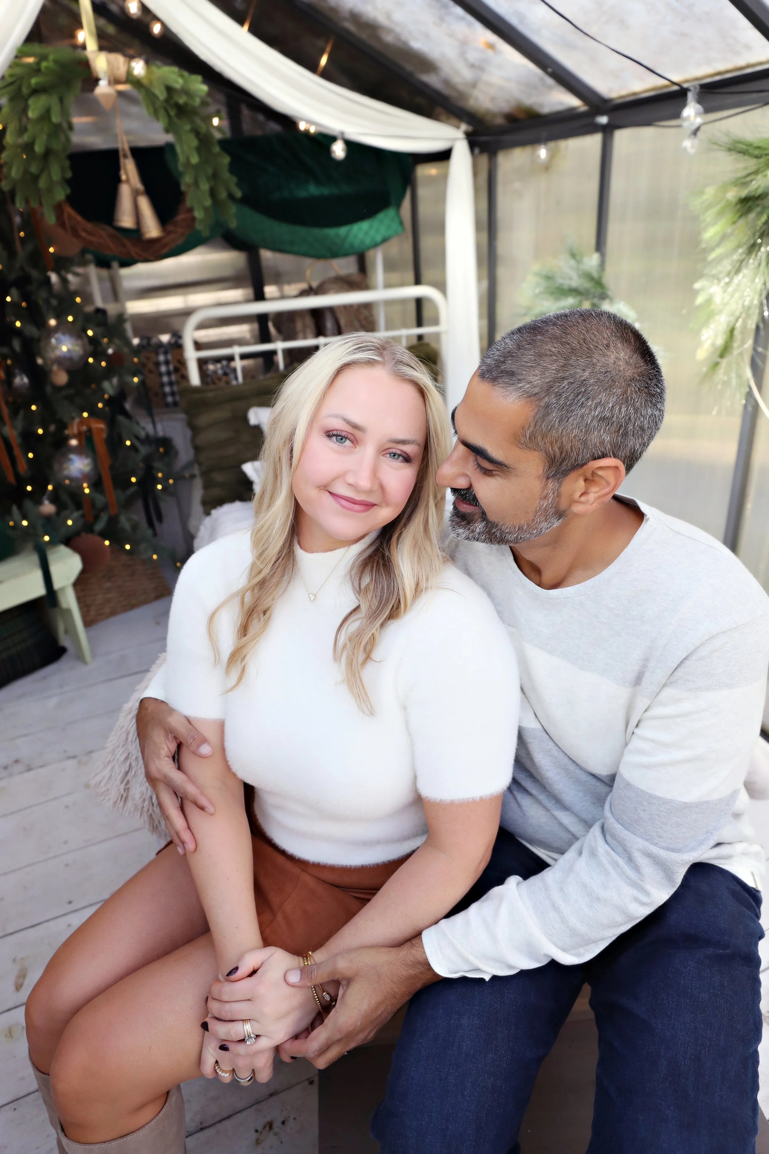 A couple sitting close together near a decorated Christmas tree, inside a cozy, decorated space with string lights, plants, and festive ornaments.