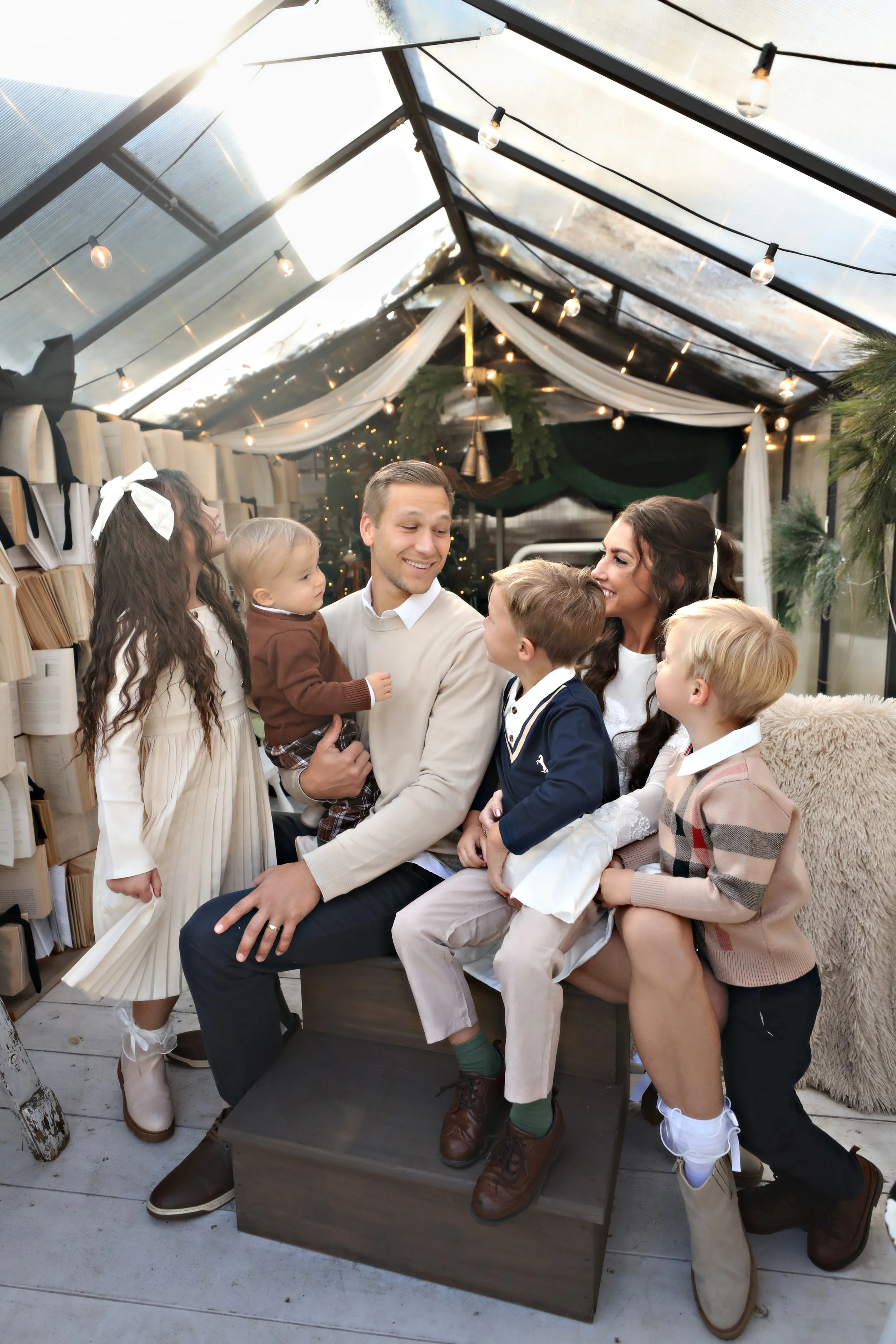 Family gathered in a cozy, decorated glass greenhouse, sharing joyful moments during the holidays with a Christmas tree, string lights, and festive decor.