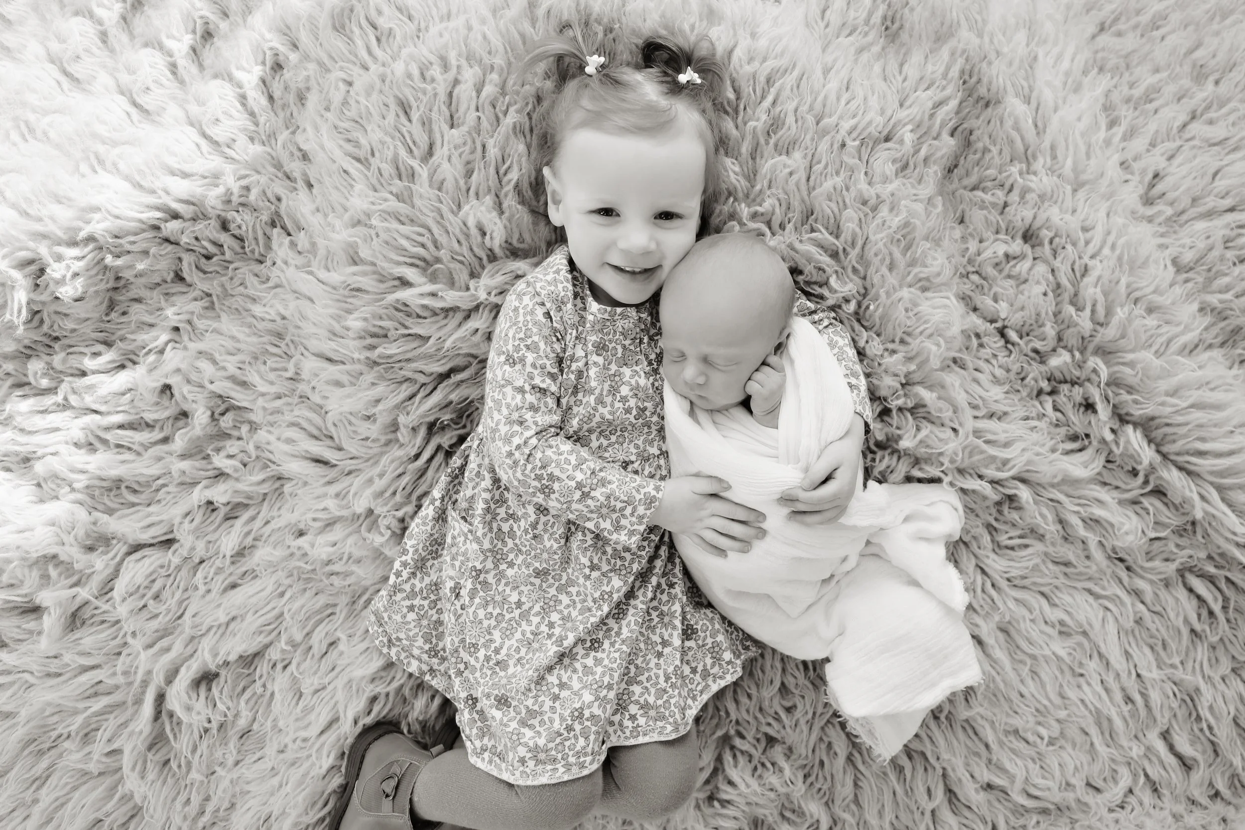 A young girl lying on a fluffy rug, holding a newborn wrapped in a blanket, both smiling and looking at the camera.
