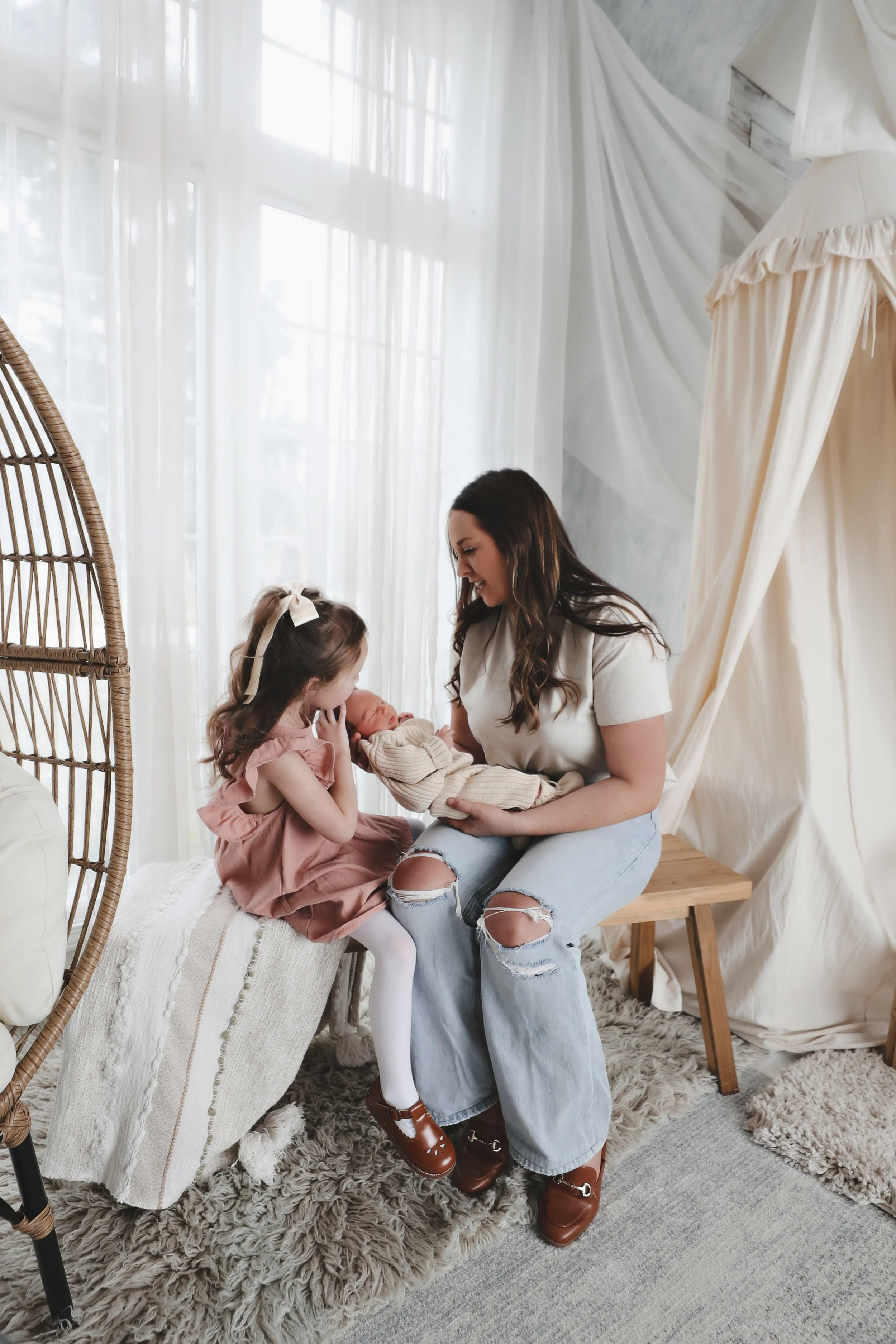 A woman holding a newborn baby, a young girl sitting beside her, all smiling inside a cozy, well-lit room with white curtains and a beige bean bag