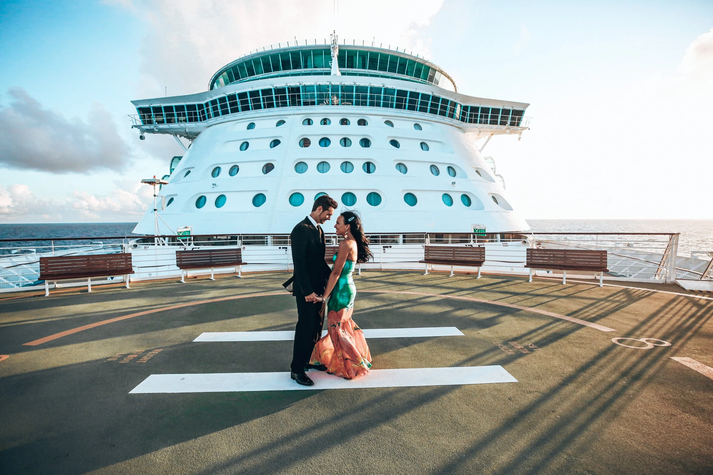 A couple dressed in formal attire standing on a ship's helipad, holding hands and looking at each other, with a large cruise ship in the background and the ocean extending to the horizon.