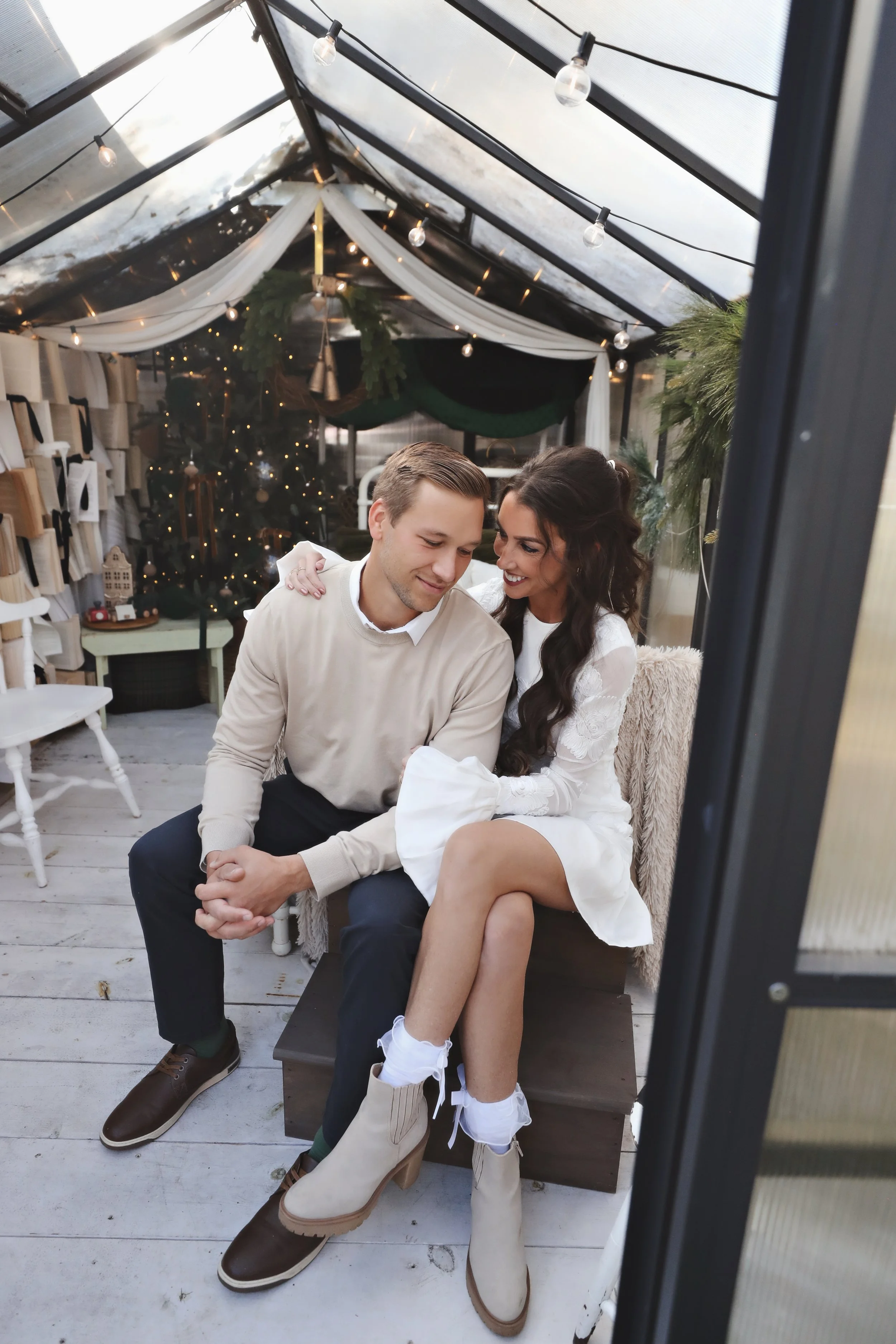 A couple sitting close together inside a decorated greenhouse, smiling and embracing, with a Christmas tree and holiday decorations in the background.