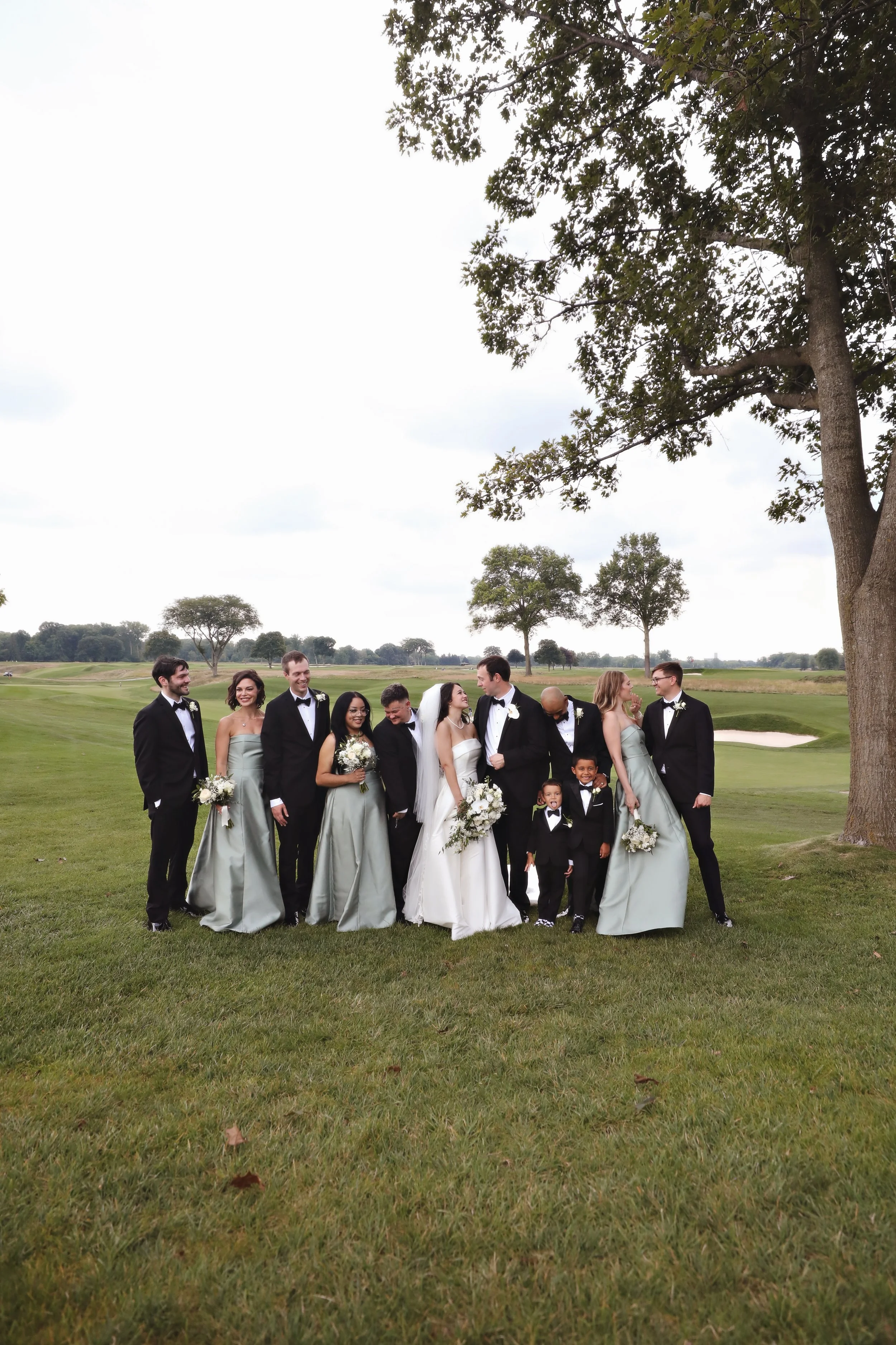 Group of wedding guests in formal attire standing on a grassy field outdoors, with trees and a cloudy sky in the background.
