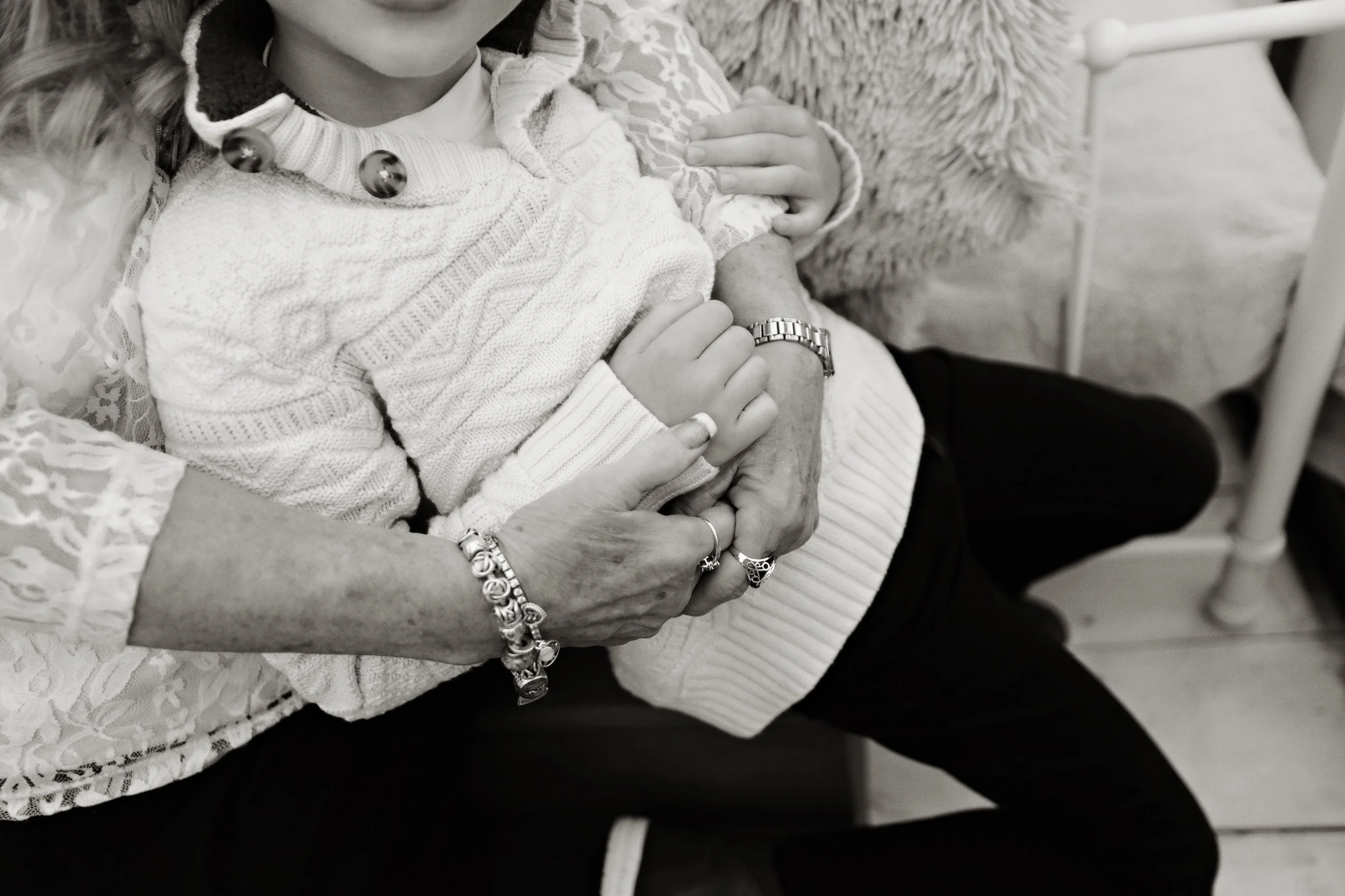 Close-up of two people holding hands, one appears to be an elderly woman and the other a younger girl. The elderly woman wears jewelry and the girl is dressed in a sweater with large buttons. They sit together on a chair, with part of a lace cloth vi