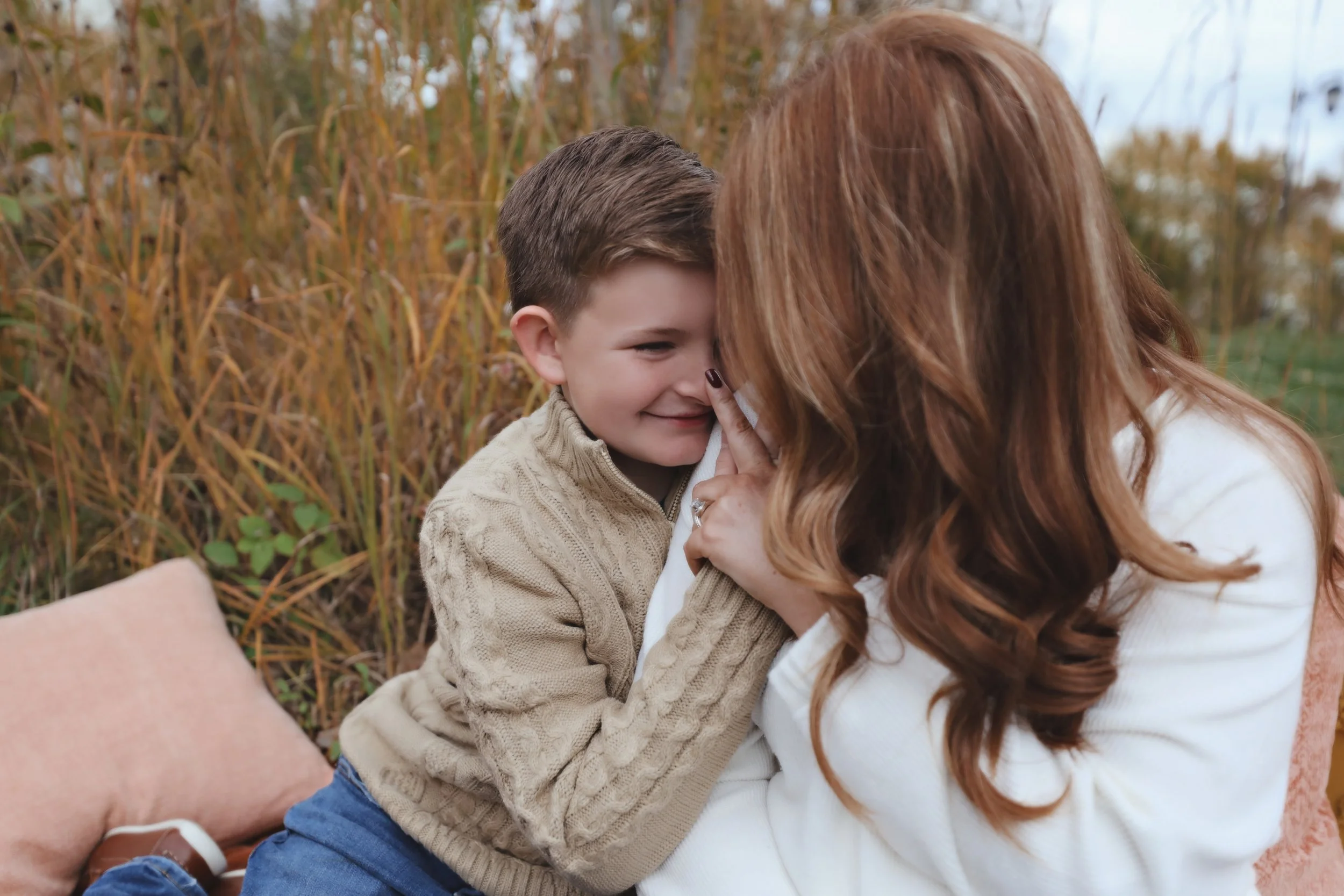 A woman with long red hair is holding a young boy with short brown hair close. They are outdoors in a field of tall autumn-colored grass. The woman is gently touching the boy's nose with her finger, and they are sharing a tender moment, smiling at ea