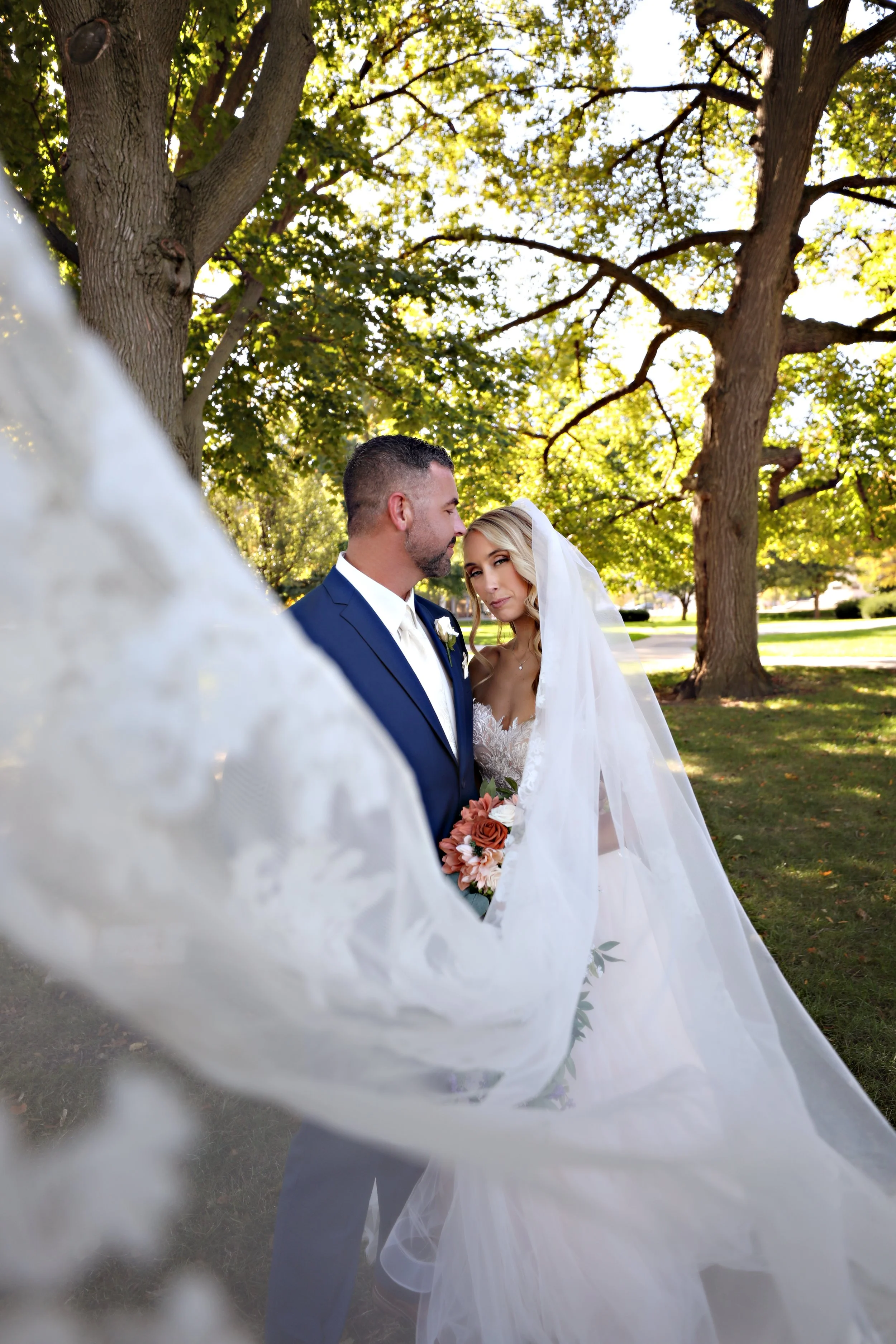 A bride and groom during their outdoor wedding, with the bride holding a bouquet of flowers, standing under large trees on a sunny day.
