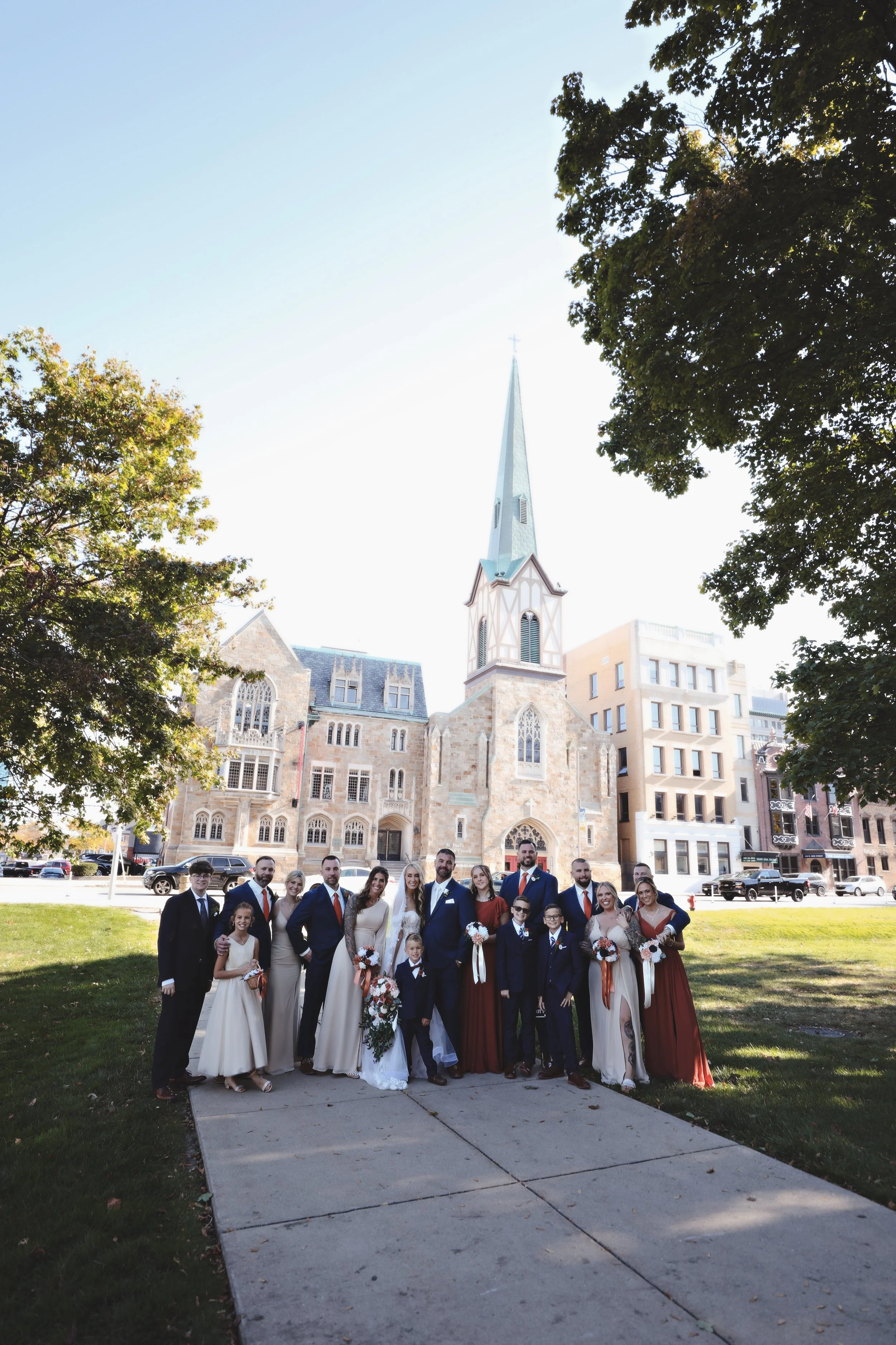 Group of people dressed for a wedding standing outside a church with a tall steeple, trees, and buildings in the background.