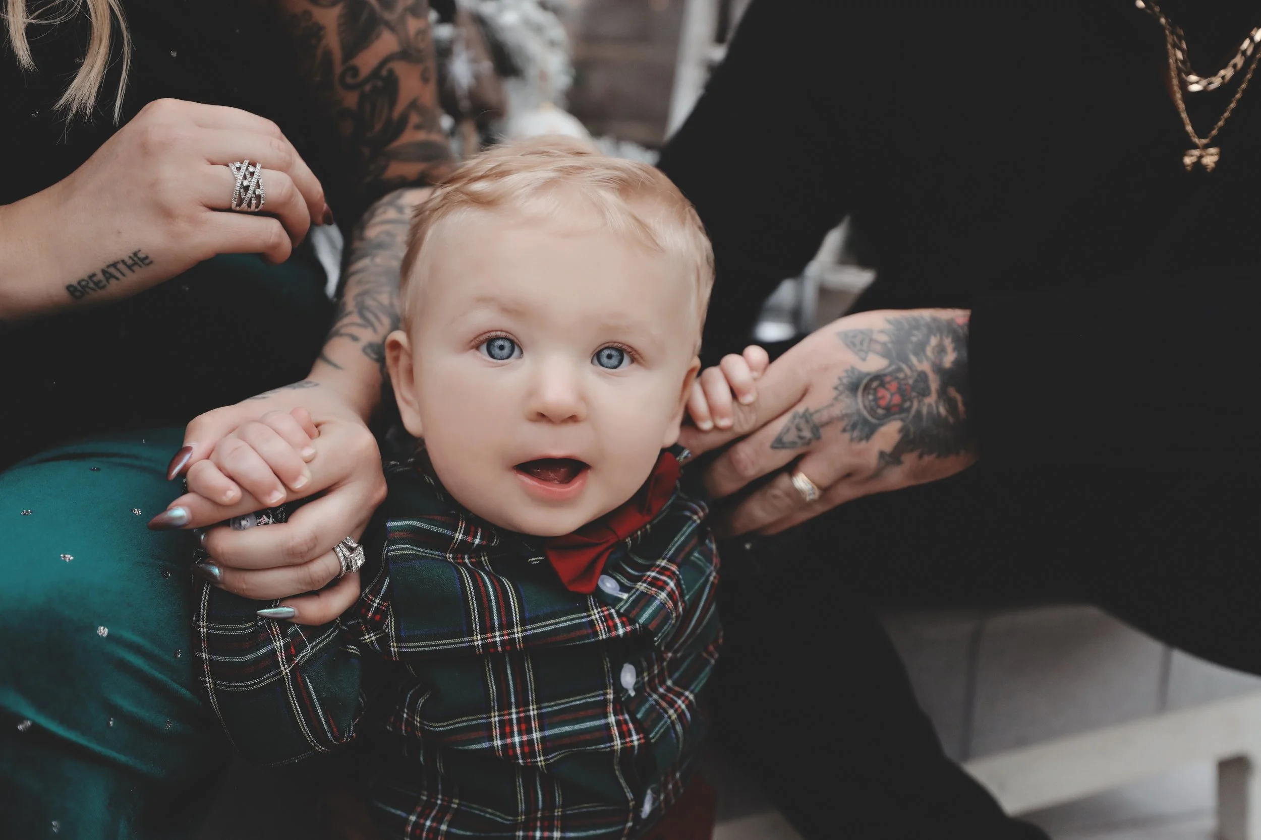 A young child with blonde hair and blue eyes wearing a plaid shirt and red bowtie, looking at the camera while being held by adults with tattoos on their arms.