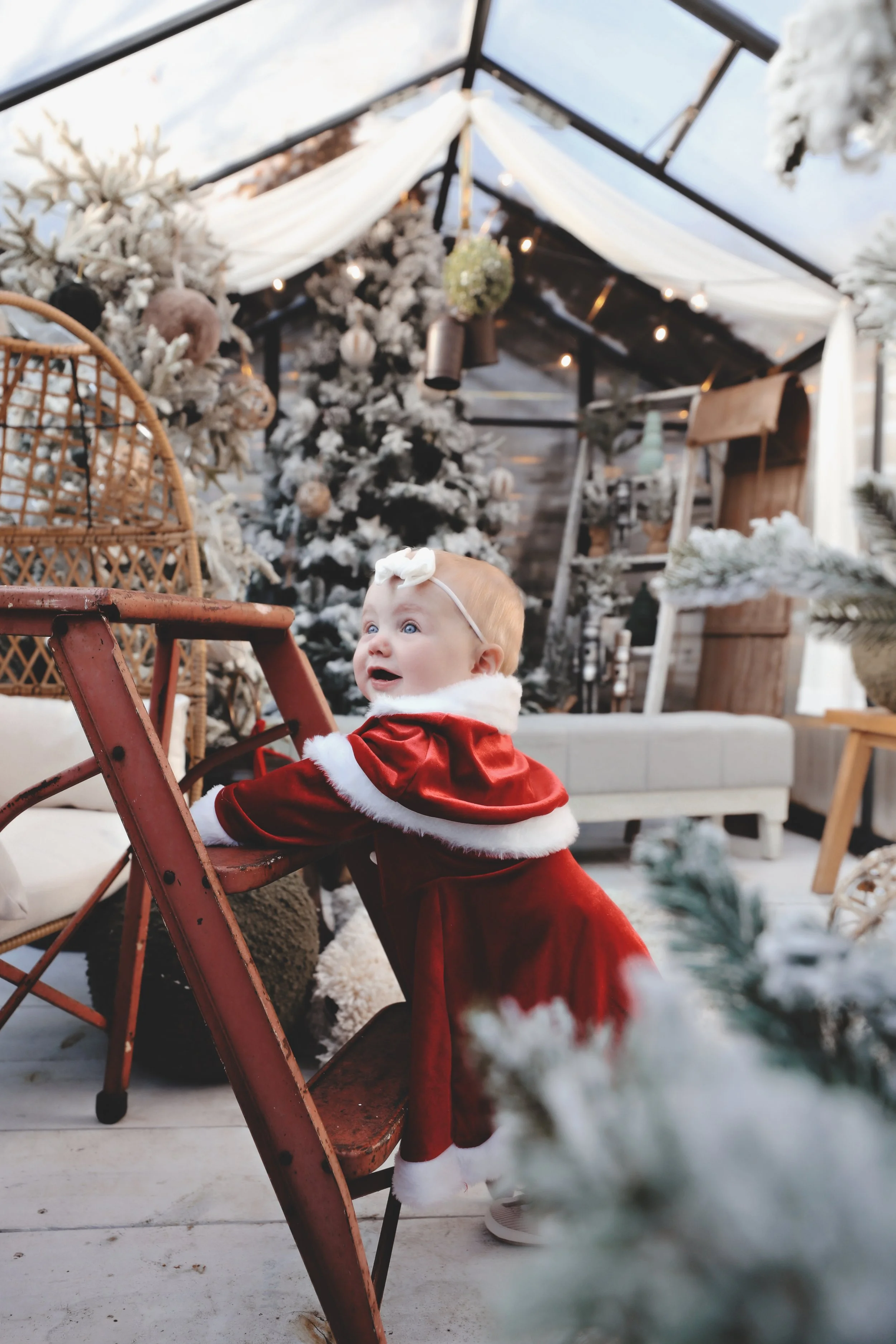 A young child dressed in a red holiday outfit with white fur trim, holding onto a rust-colored chair in a decorated, snowy-themed indoor space with Christmas trees and festive decor.