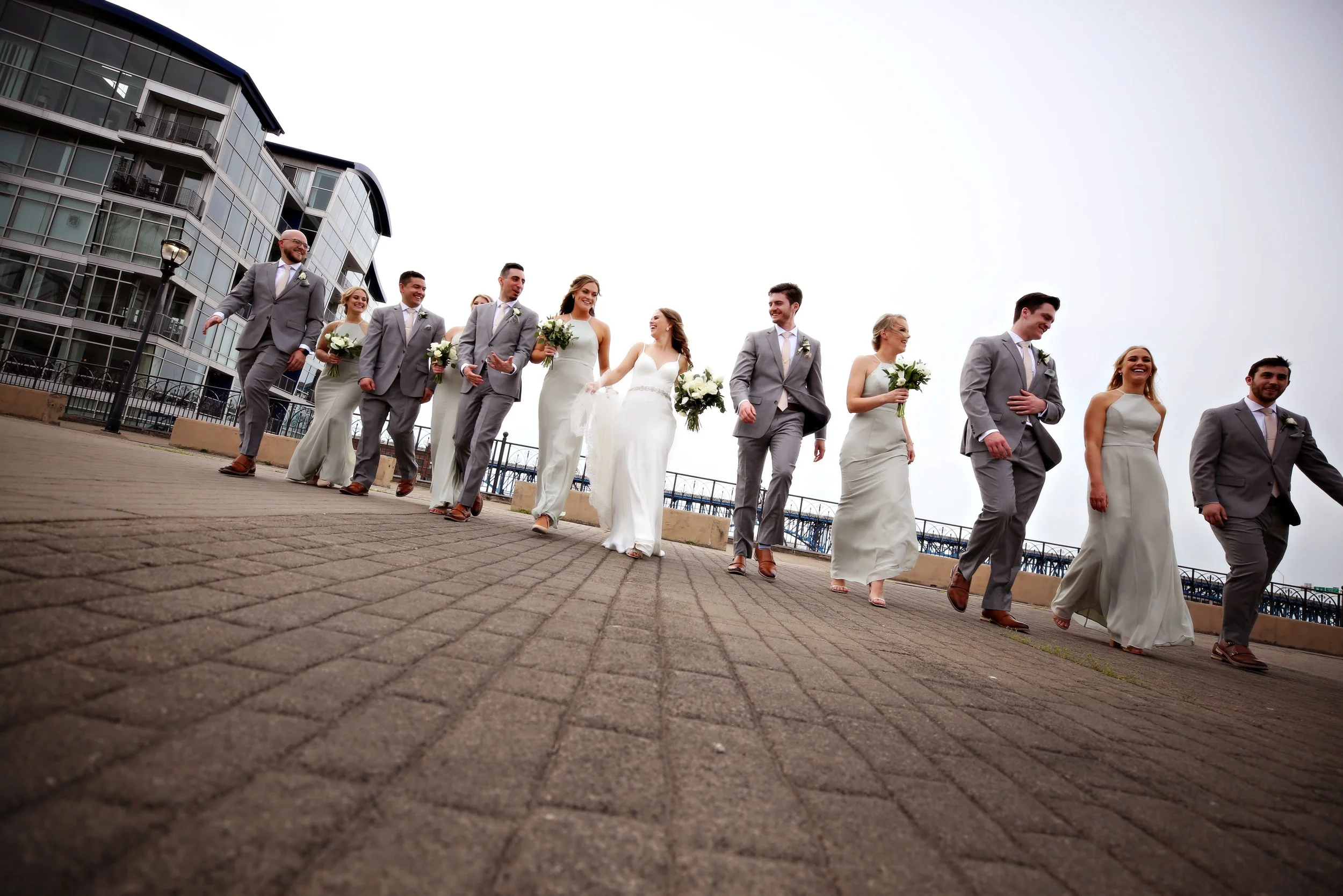 Bride and groom with bridesmaids and groomsmen walking outdoors on a paved area near modern buildings, dressed in formal wedding attire, holding bouquets and smiling.