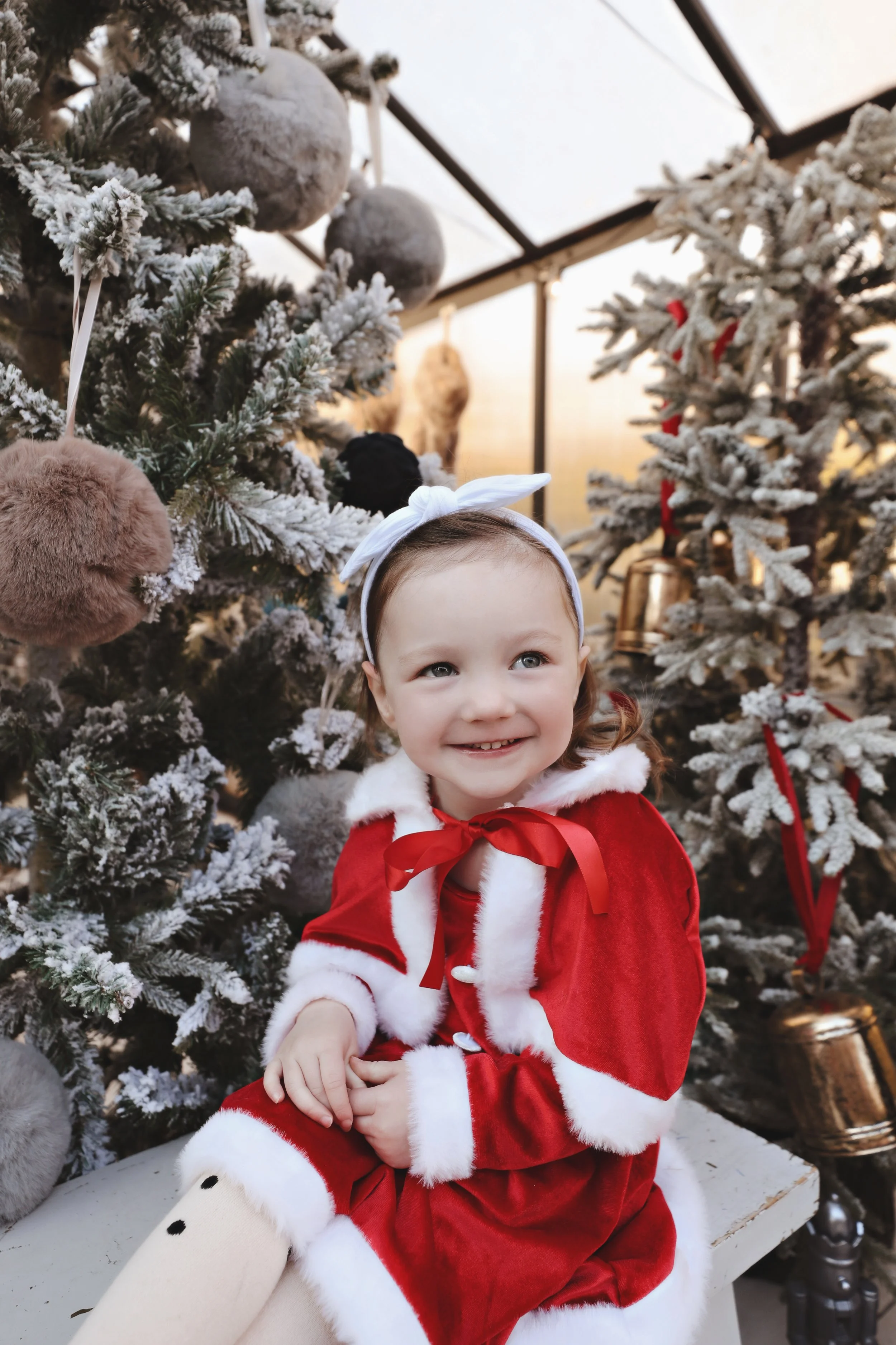 A smiling young girl dressed in a red and white Santa outfit, sitting in front of Christmas decorated trees with ornaments, in a festive setting.