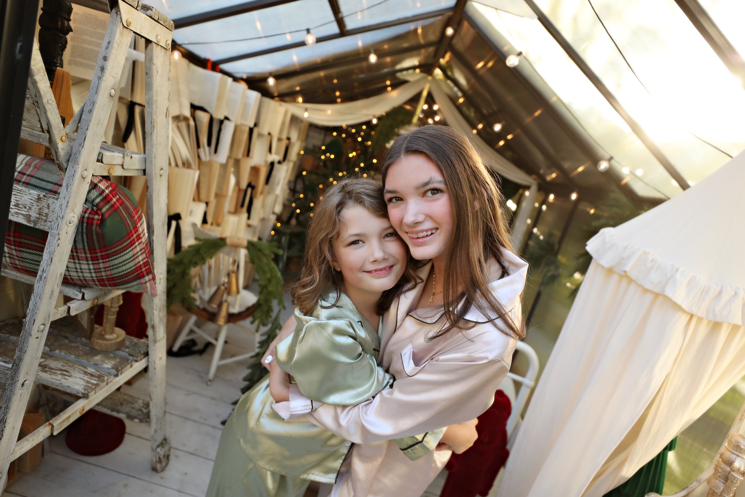 A woman and a young girl happily hugging each other indoors decorated with string lights, Christmas greenery, and holiday decor during the daytime.