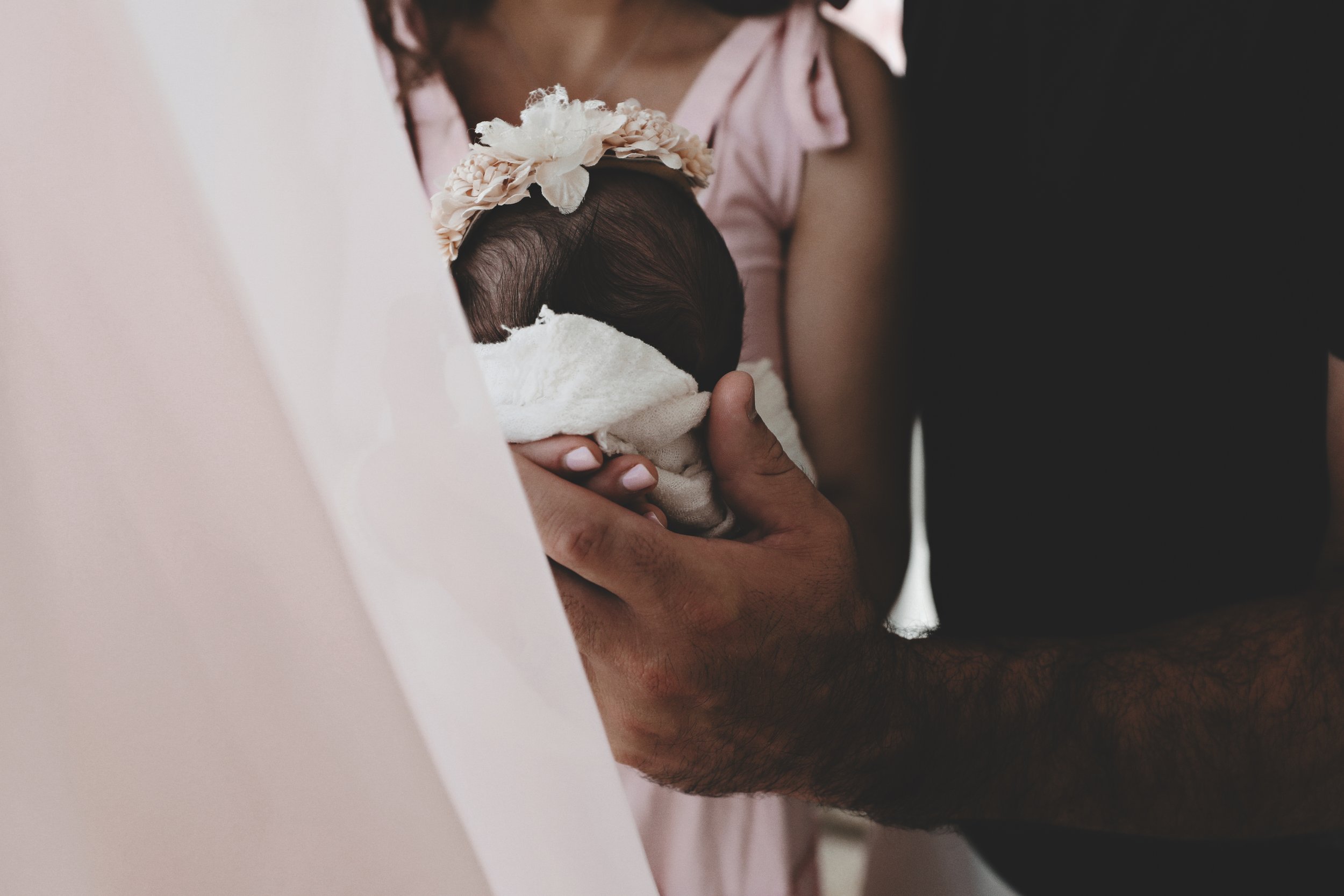 A person holding a newborn baby wrapped in a white blanket, wearing a floral headband, with another person supporting from the side.