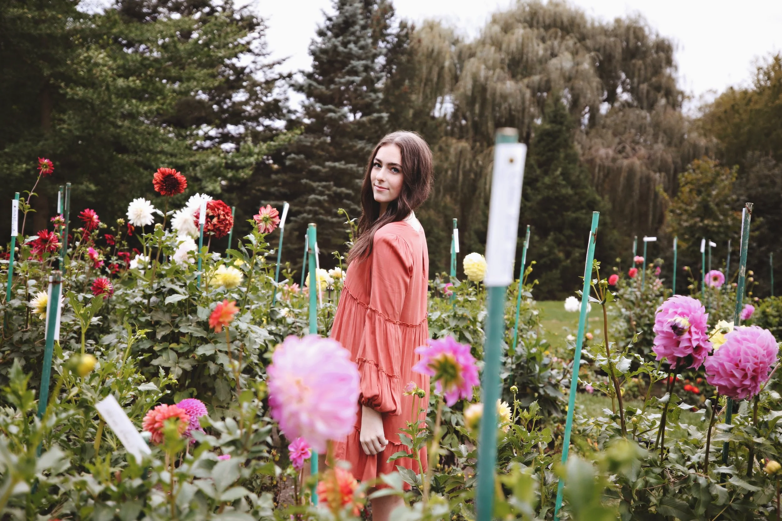 A young woman in a coral dress standing among blooming dahlias in a garden with green trees and a cloudy sky in the background.