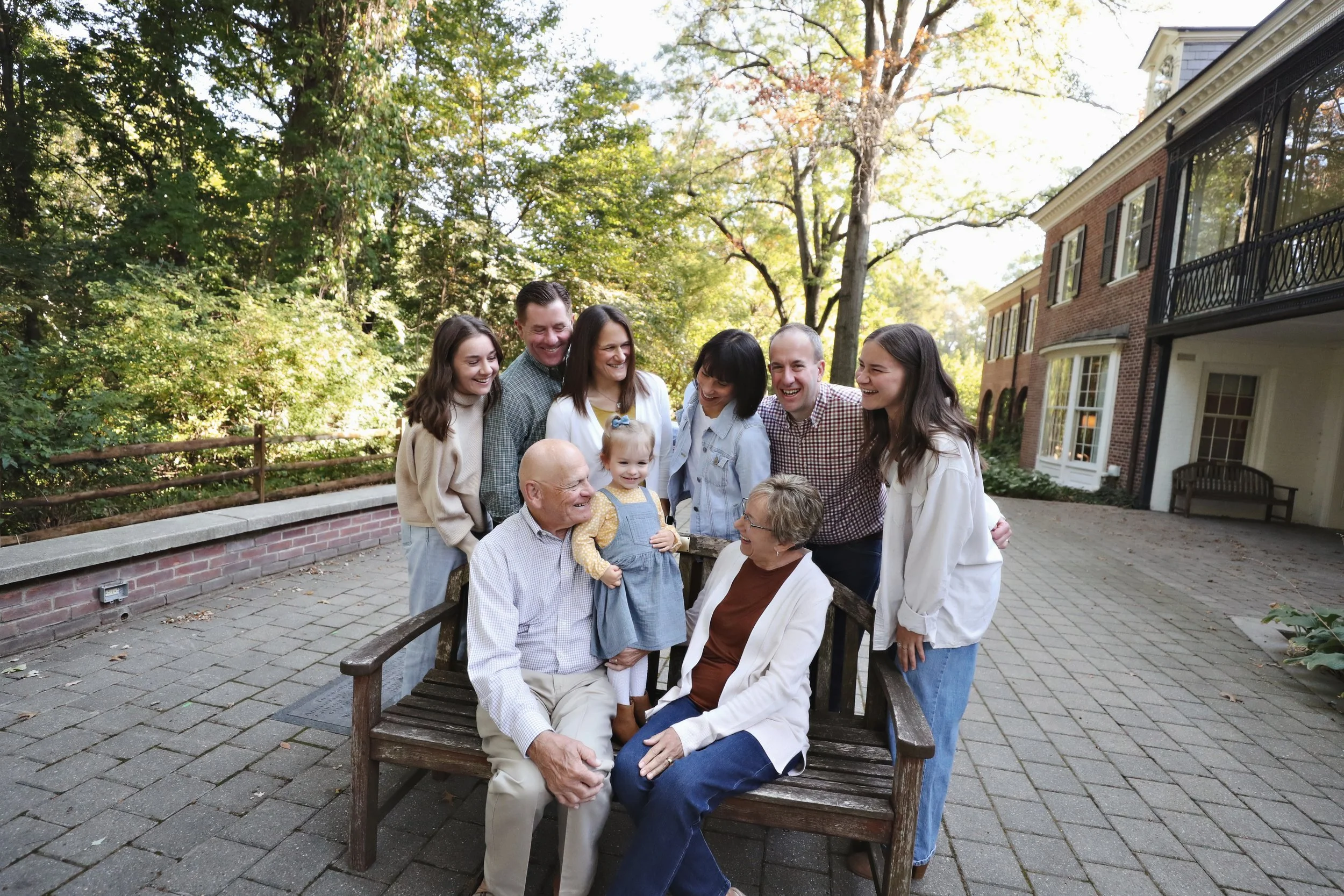 A family gathering outdoors on a bench, with multiple generations, including elderly grandparents, middle-aged adults, teenagers, and a young girl, all smiling and enjoying each other's company in a courtyard area with trees and residential buildings