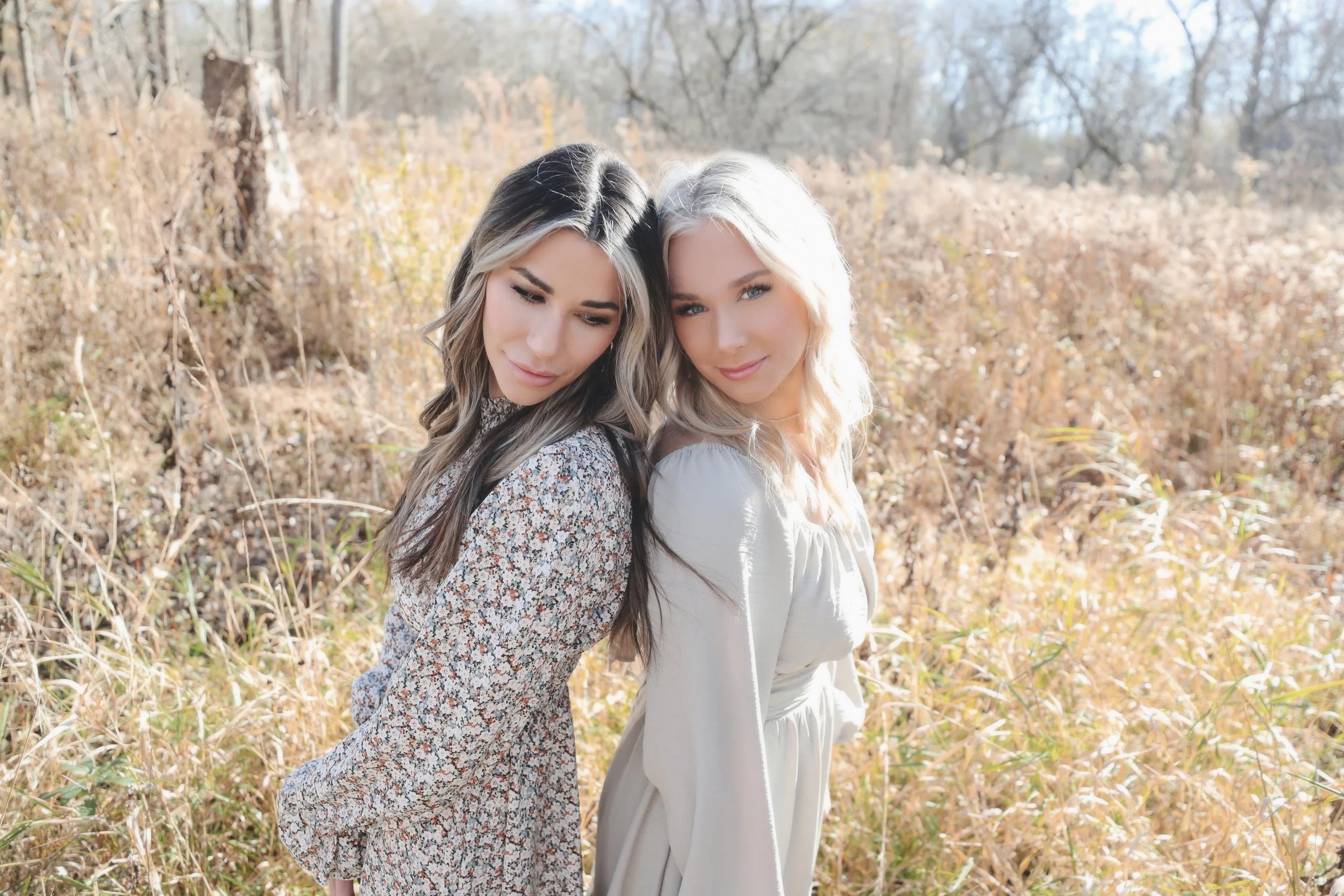 Two young women stand close together in a field of tall, dry grass and weeds with trees in the background, both looking downward with slight smiles, dressed in long sleeves and light-colored dresses.