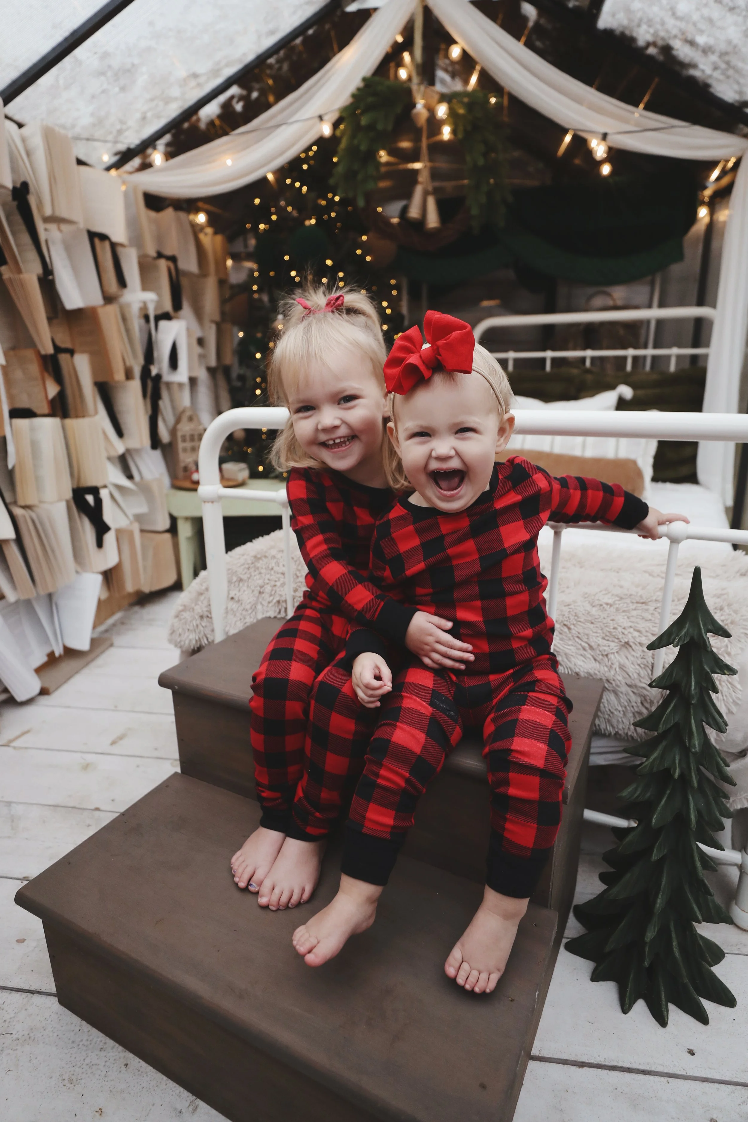 Two young girls in matching red and black plaid pajamas sitting on a step, smiling and laughing in a festive, decorated room with Christmas trees and lights.
