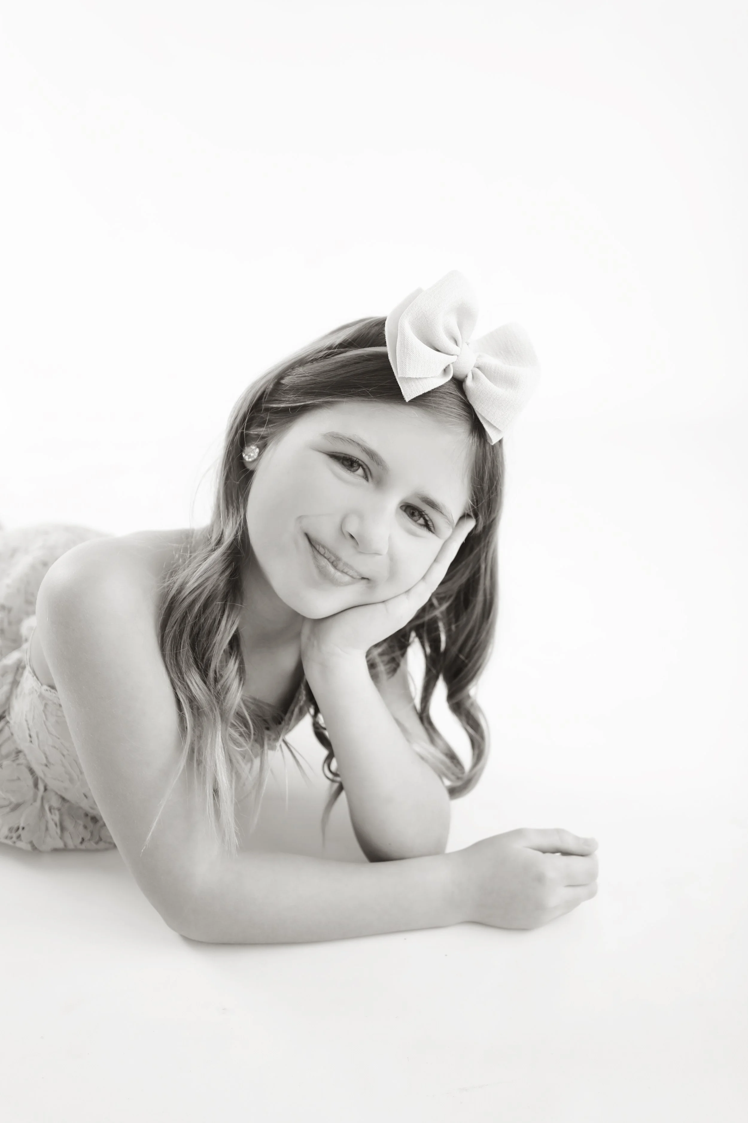 Black and white portrait of a young girl lying on her side, resting her head on her hand, with a bow in her hair, smiling gently at the camera.