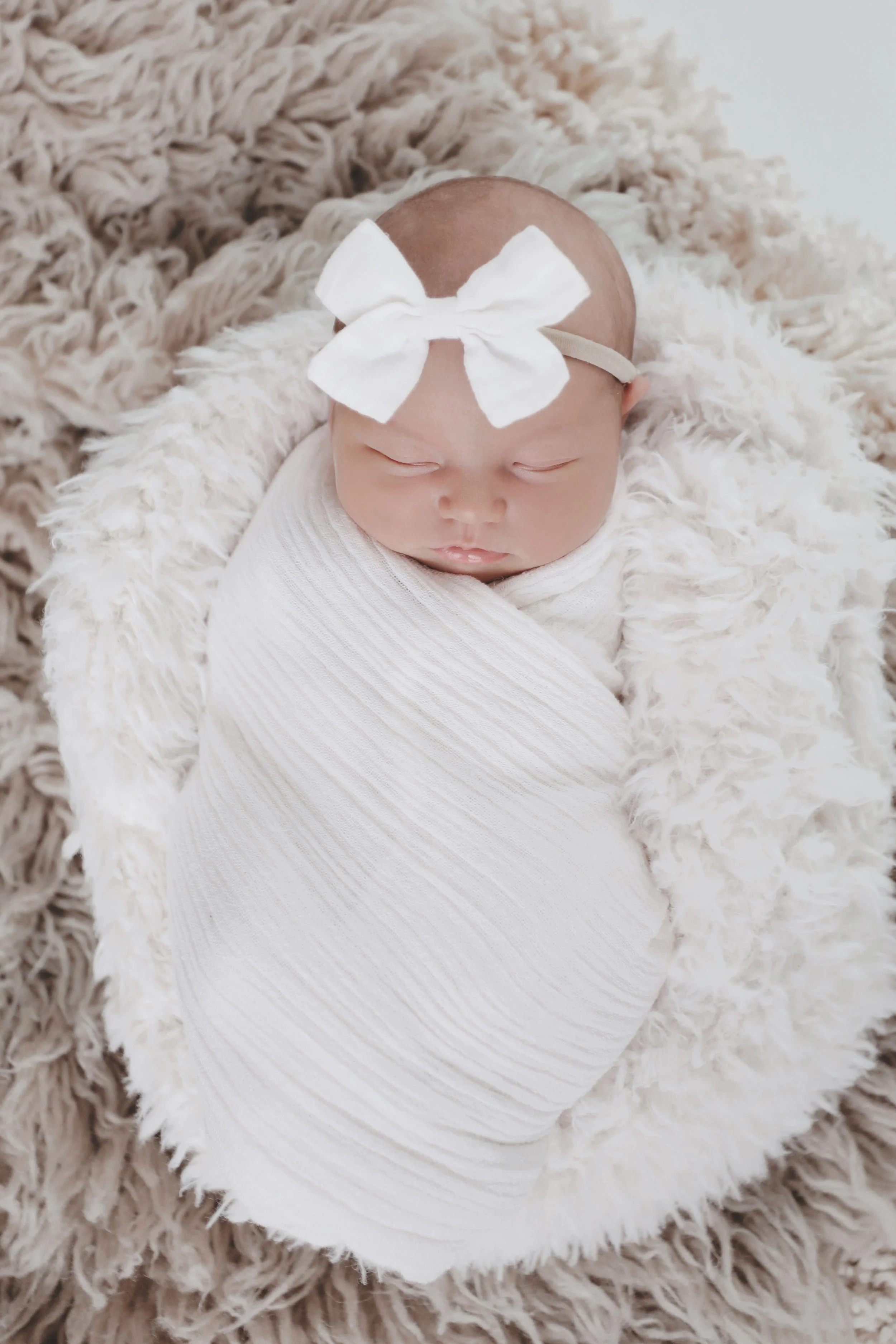 A sleeping newborn baby swaddled in a white blanket, wearing a large white bow headband, resting on a fluffy beige blanket.