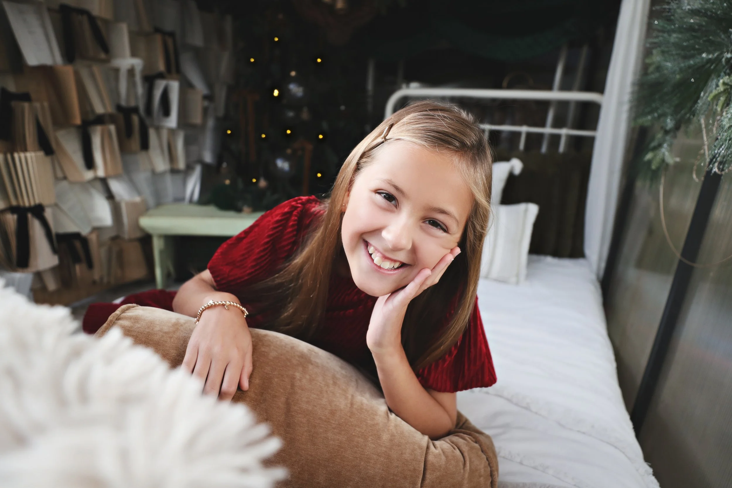 A girl with brown hair wearing a red shirt, smiling and resting her face on her hand, lying on a bed in a cozy, decorated room with a decorated Christmas tree in the background.