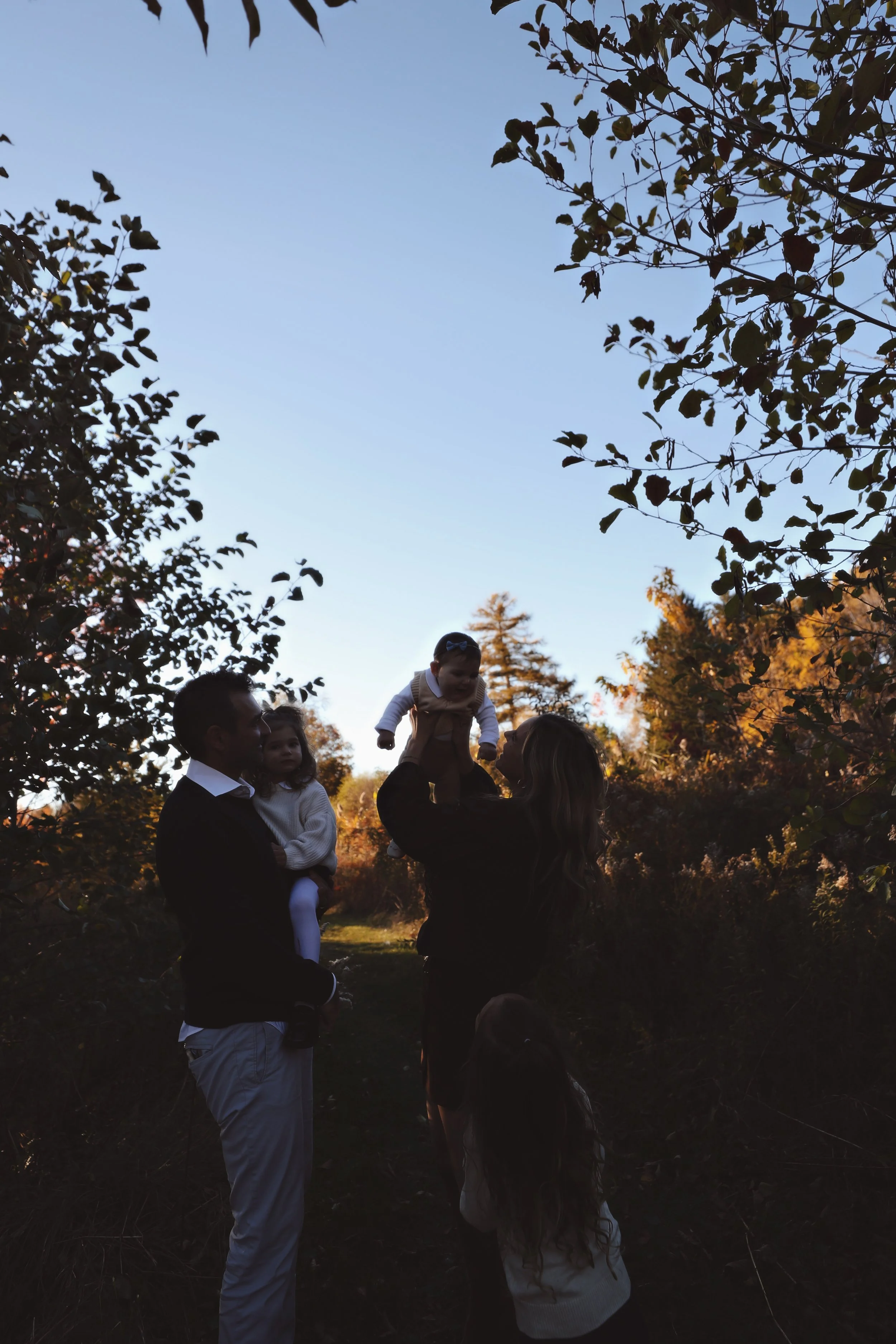 Family standing outdoors on a trail surrounded by trees, with a clear blue sky overhead. The woman is lifting a young child into the air while a man holds another young child.