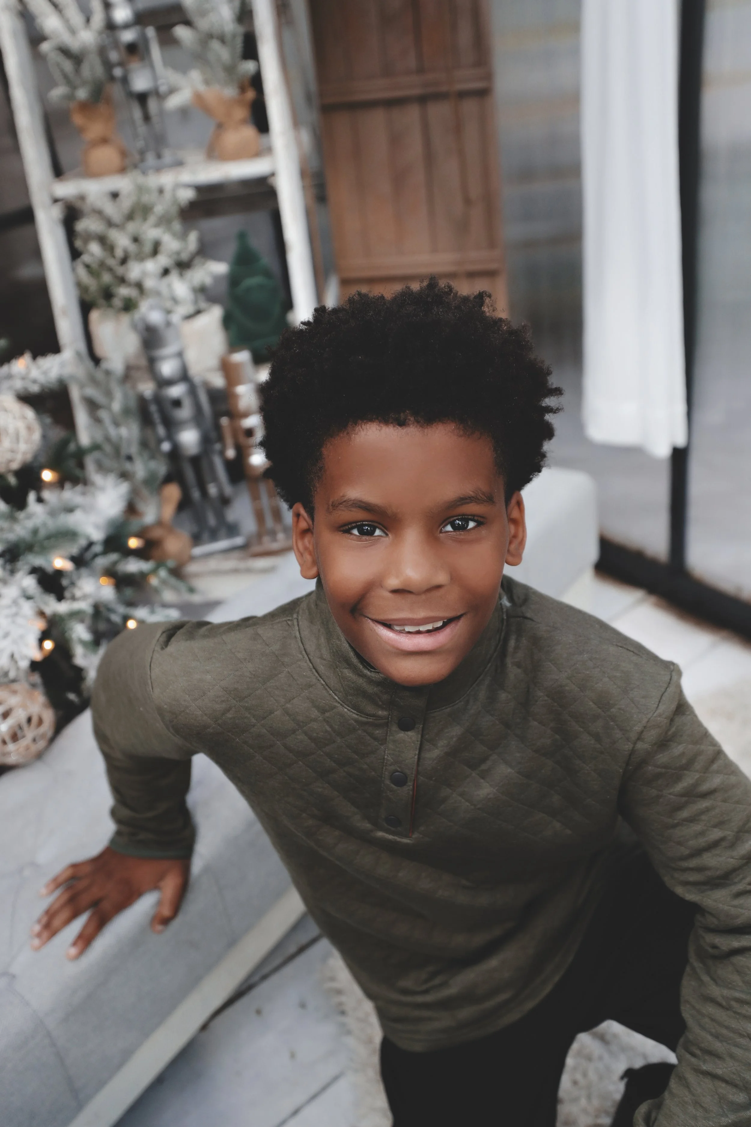 A smiling young boy with dark curly hair leaning on a gray sofa, indoors near a decorated Christmas tree and holiday decorations.