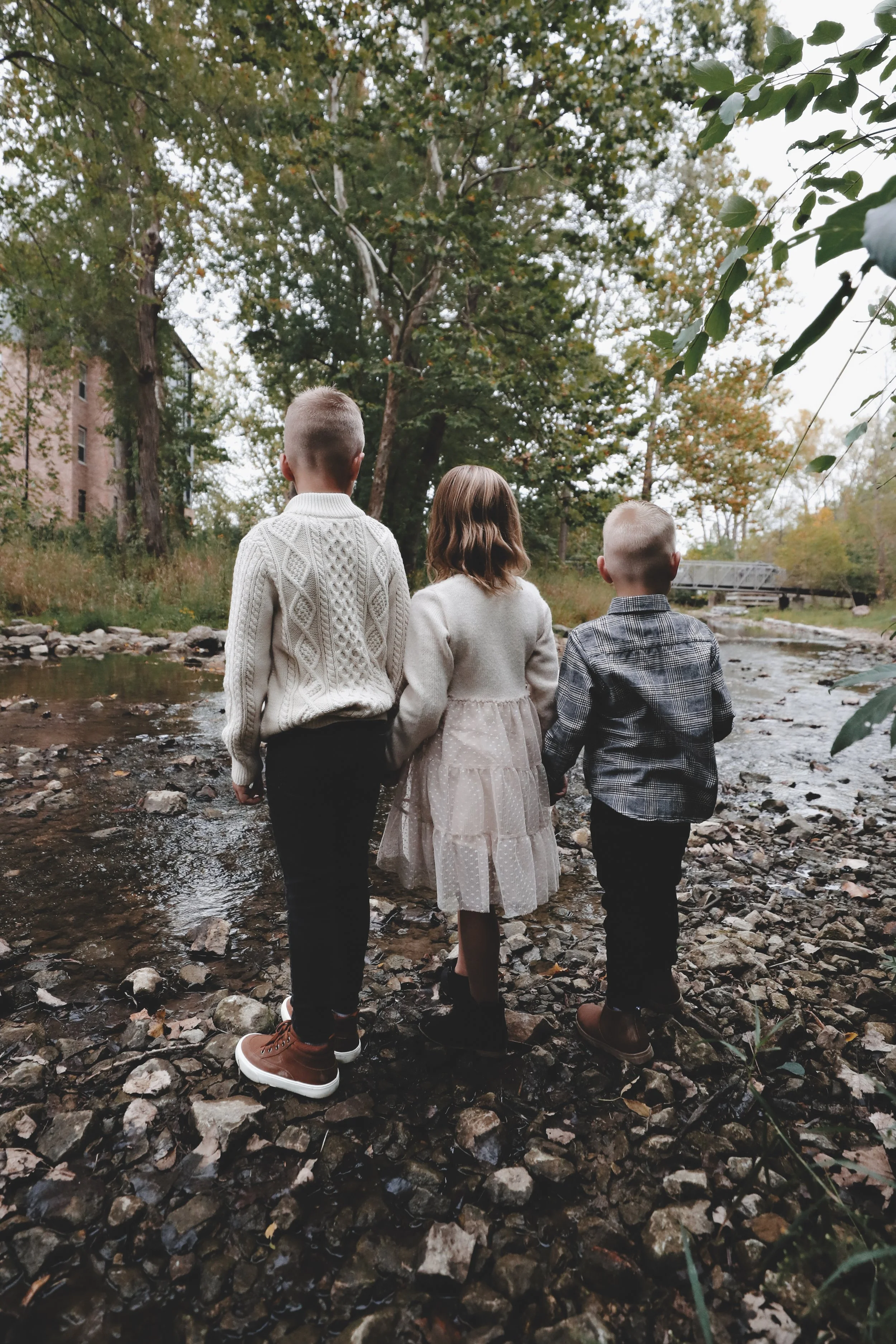 Three children, two boys and a girl, holding hands while walking through a shallow creek in a forest area during daytime.