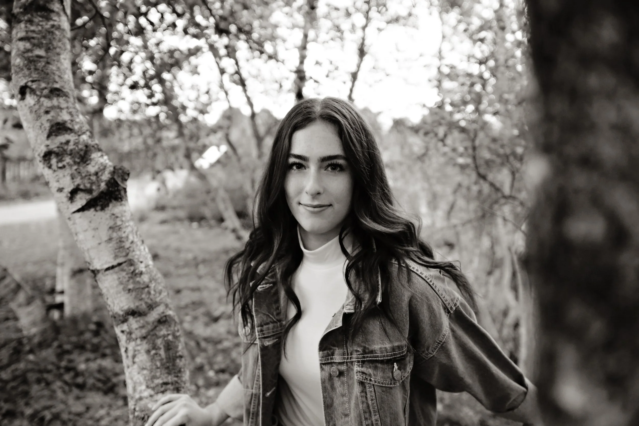 A young woman with long dark wavy hair, wearing a denim jacket over a white top, stands outdoors among trees, smiling slightly at the camera in a black and white photo.