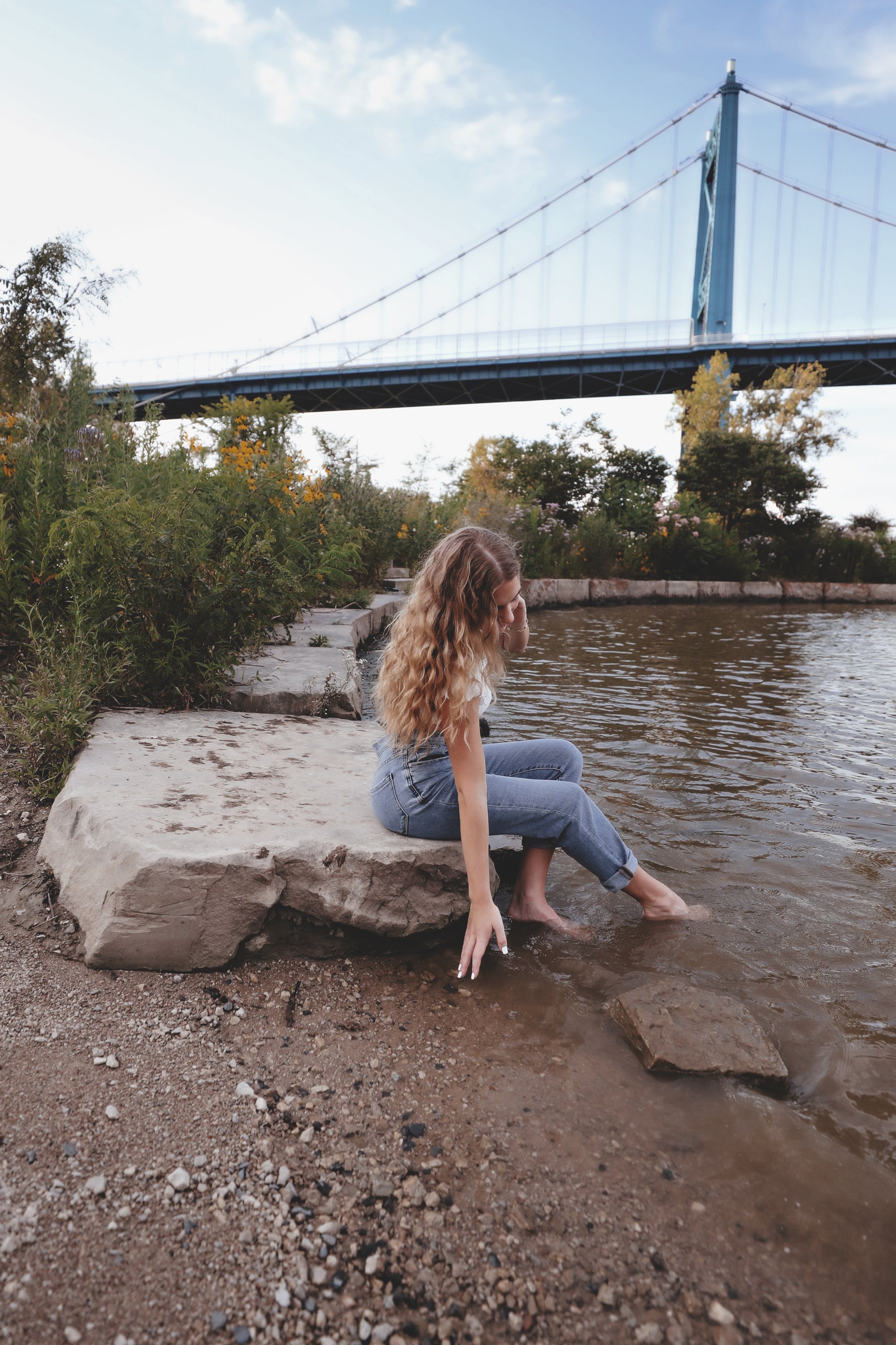 A young woman with long wavy hair sits on a large rock by a river, dipping her feet in the water, with a bridge visible in the background and surrounded by greenery.