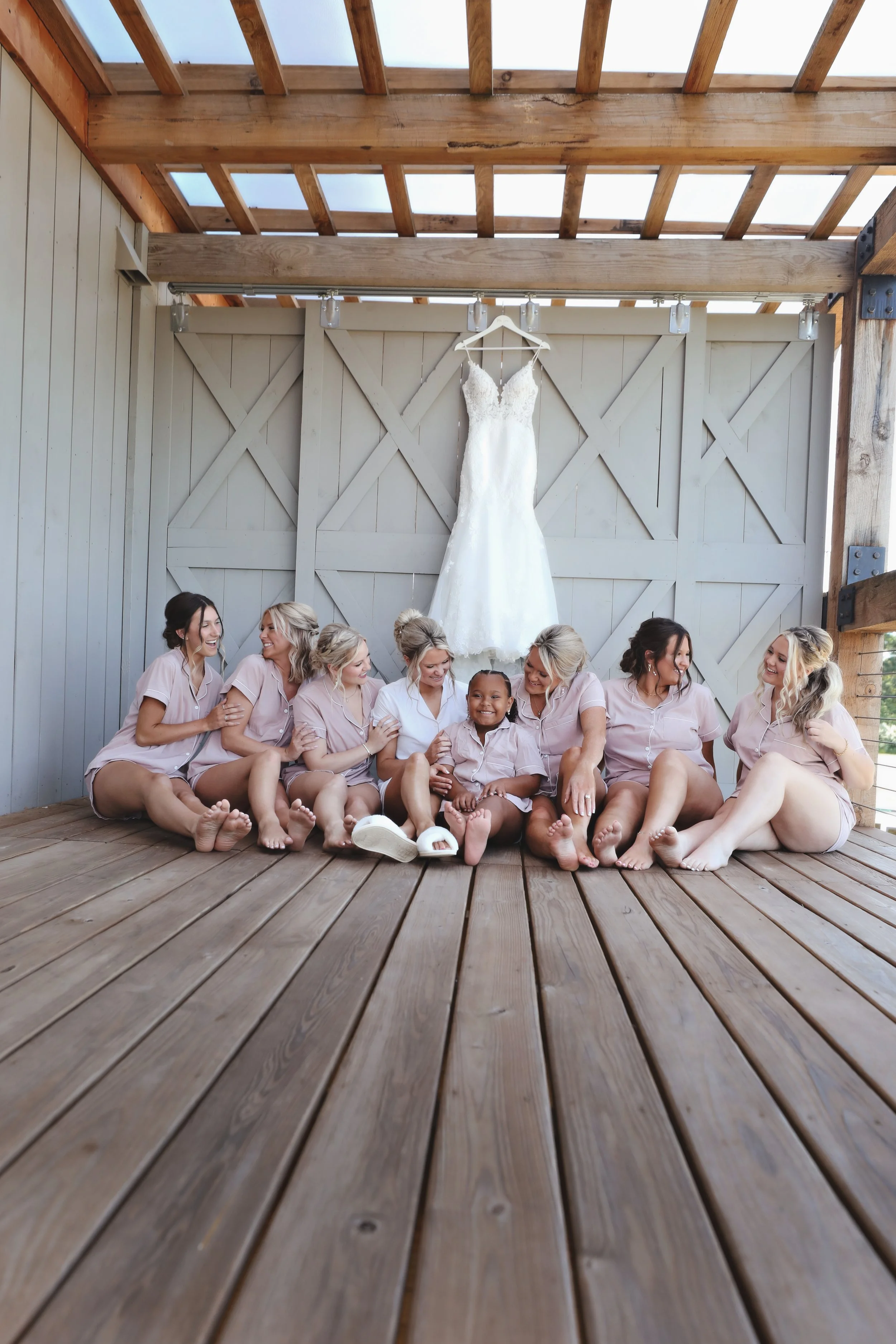Bridesmaids and a flower girl sitting on a wooden floor, smiling and laughing, in front of a white wedding dress hanging on a barn-style door.