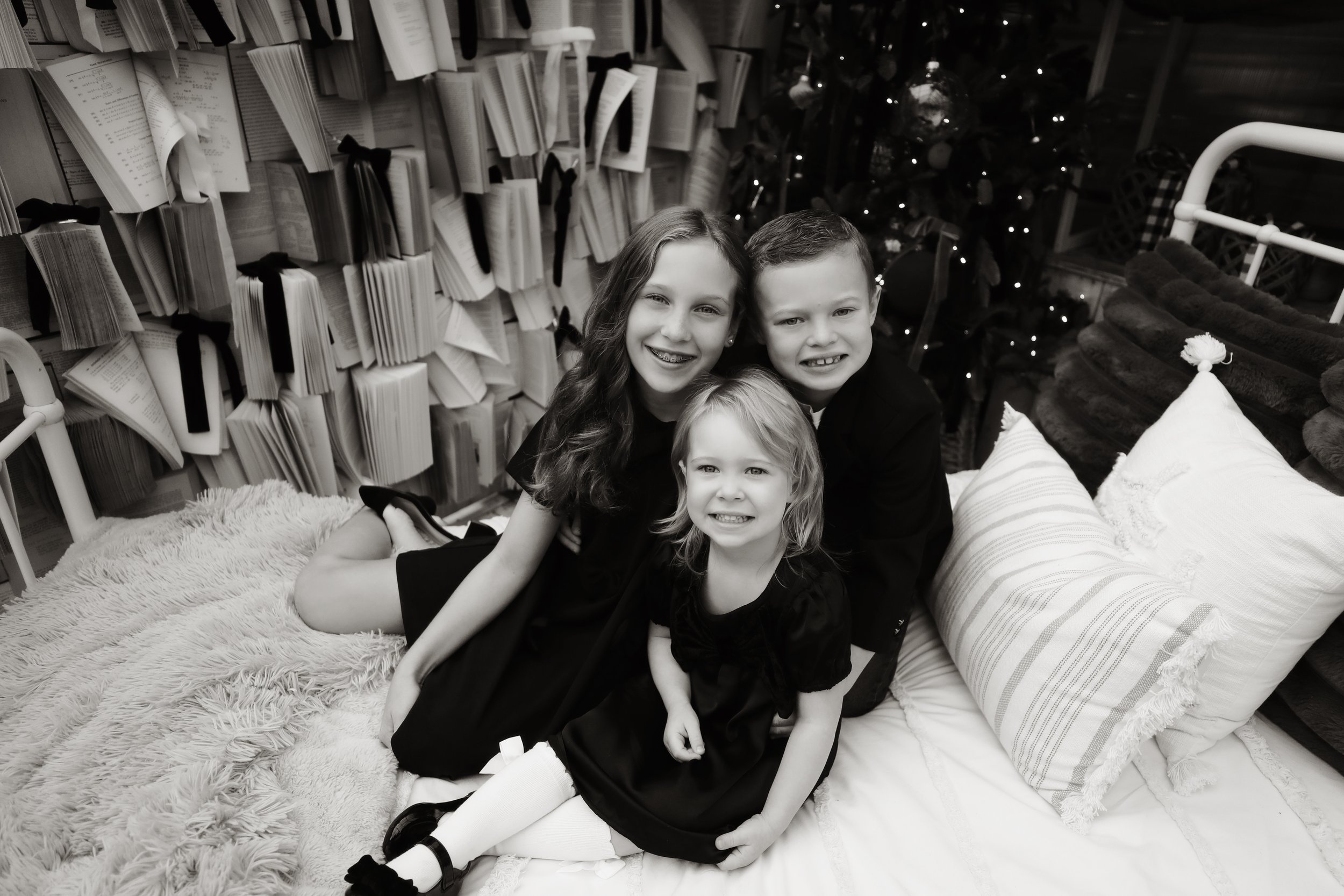 Three children, two girls and a boy, sitting on a bed with pillows, in a room decorated with Christmas lights and a decorated Christmas tree in the background, surrounded by shelves filled with books or papers.