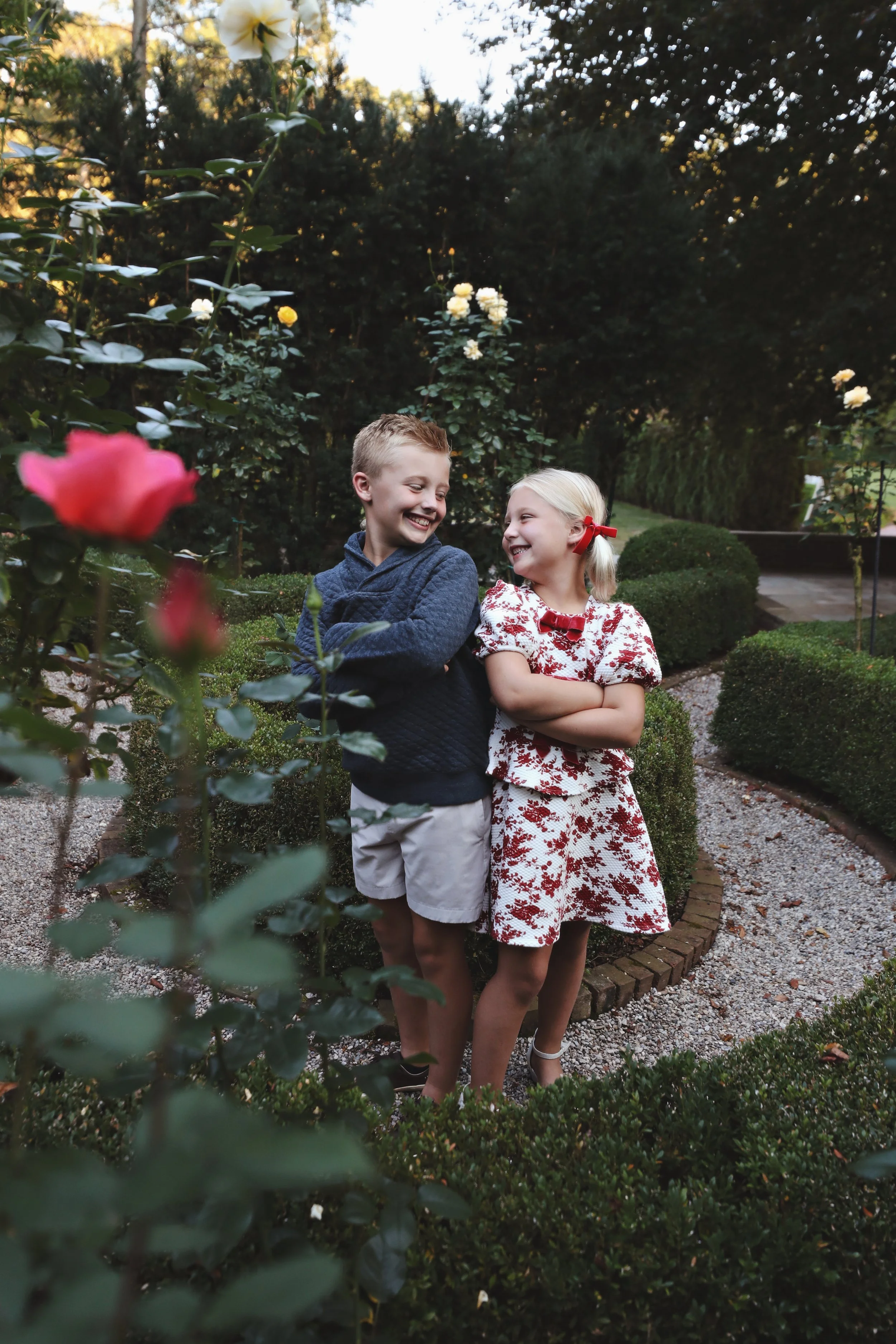 Two children, a boy and a girl, standing in a garden with bushes and flowers, smiling and looking at each other.
