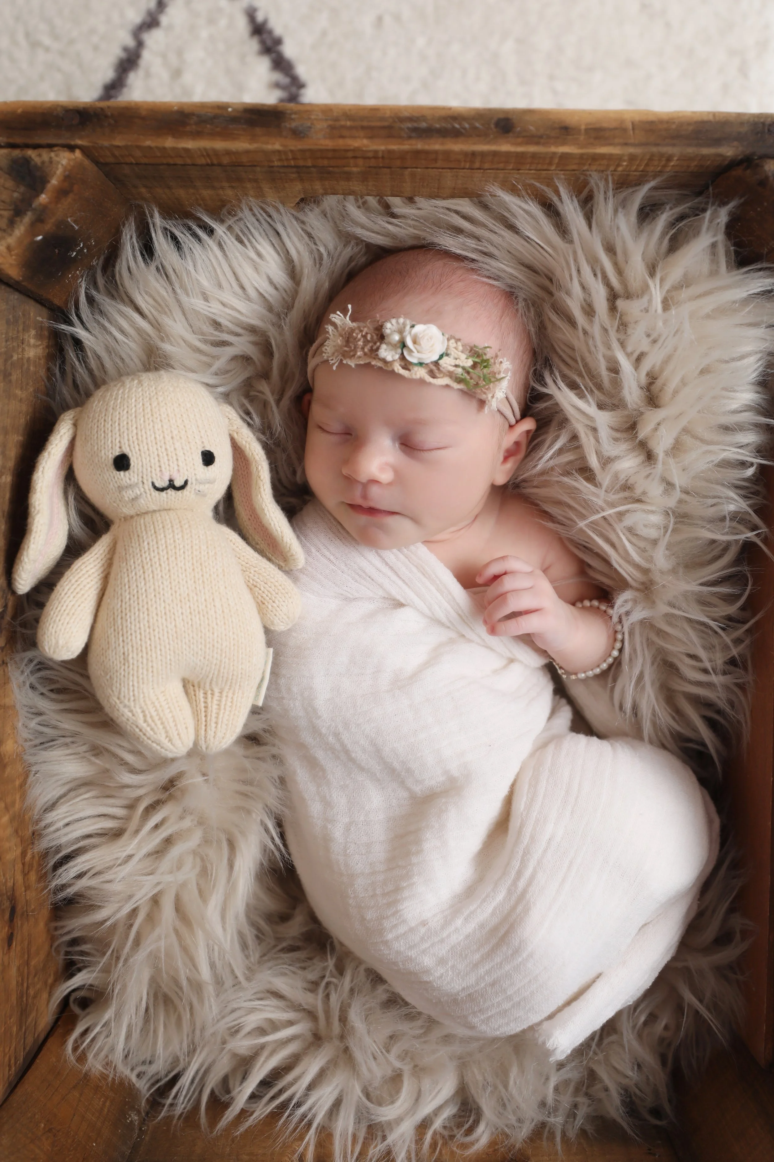 A sleeping newborn baby with a flower headband, swaddled in white, lying on a soft furry blanket inside a wooden box, next to a knitted bunny toy.