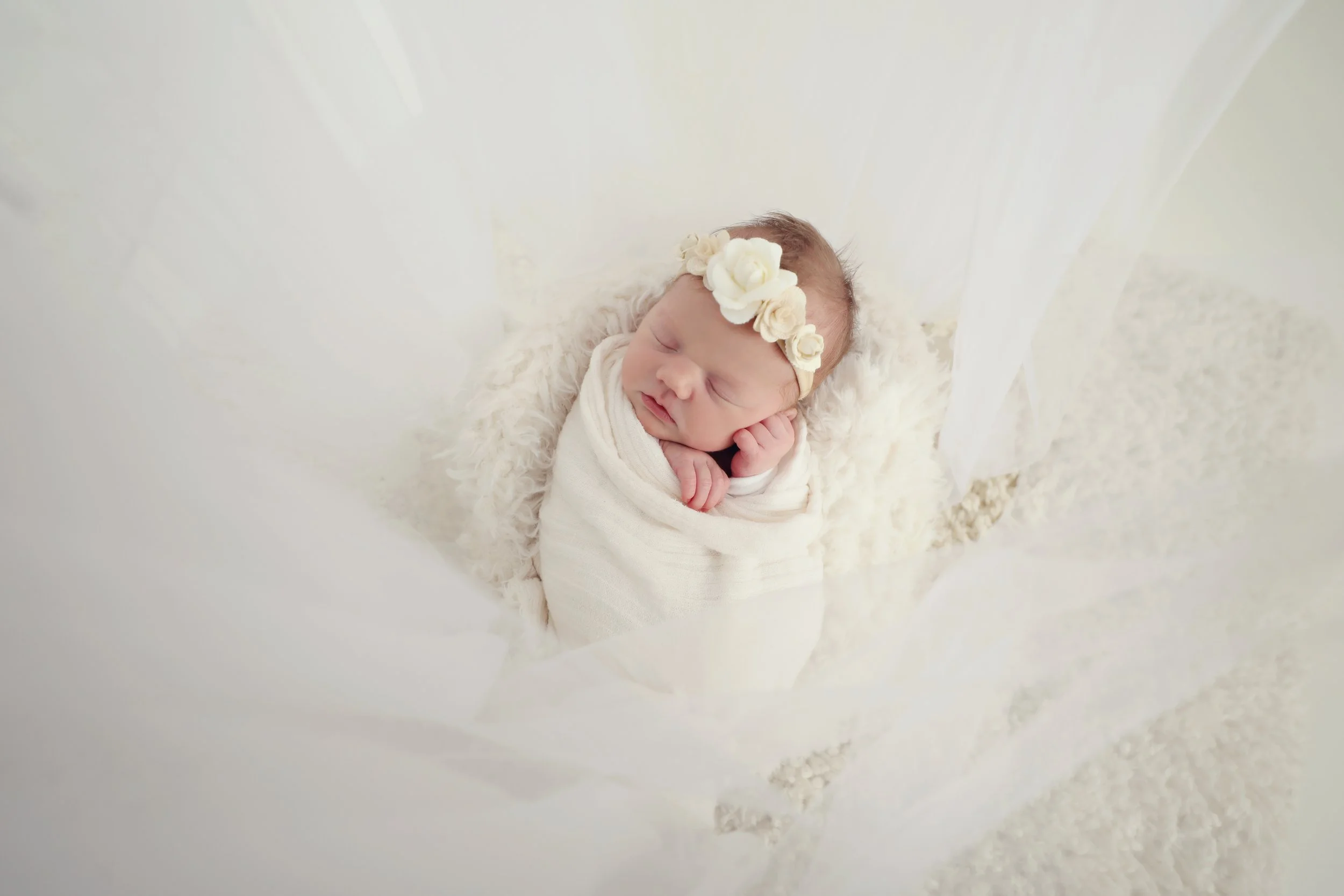 A newborn baby wrapped in a white blanket, sleeping on a soft furry surface, wearing a floral headband.