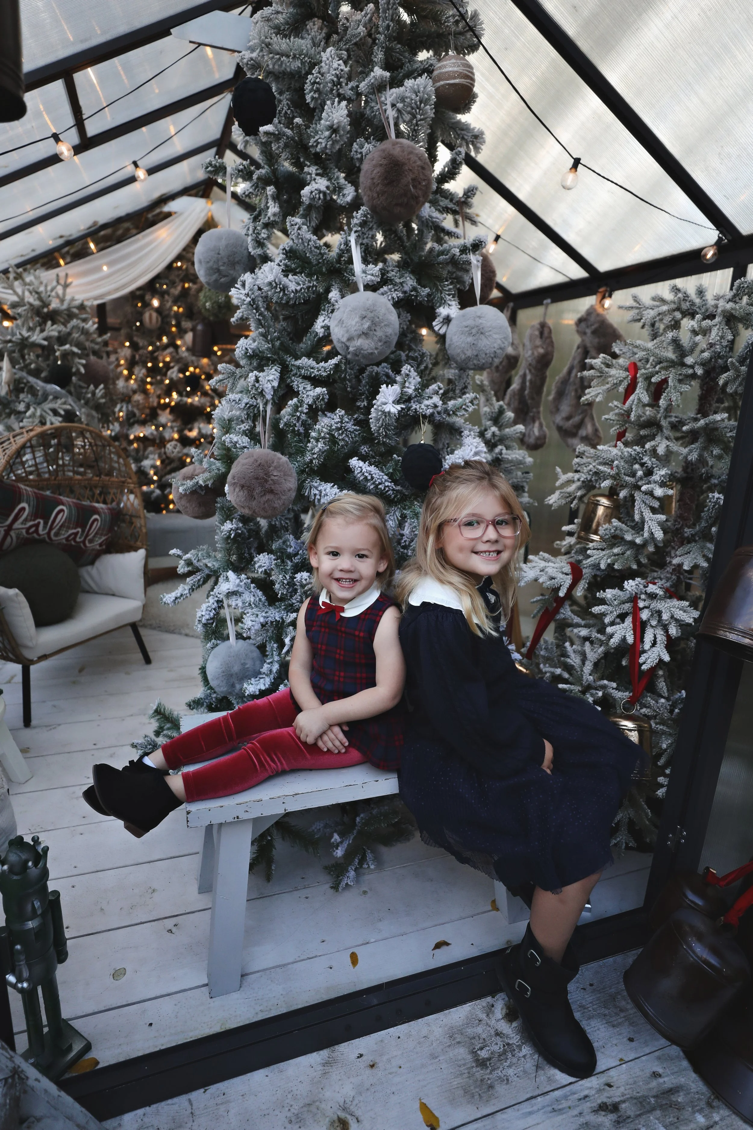 Two young girls sitting together on a white bench in front of a decorated Christmas tree with white and gray ornaments, inside a glass greenhouse. The girl on the left is wearing a plaid dress with red velvet leggings, and the girl on the right is we