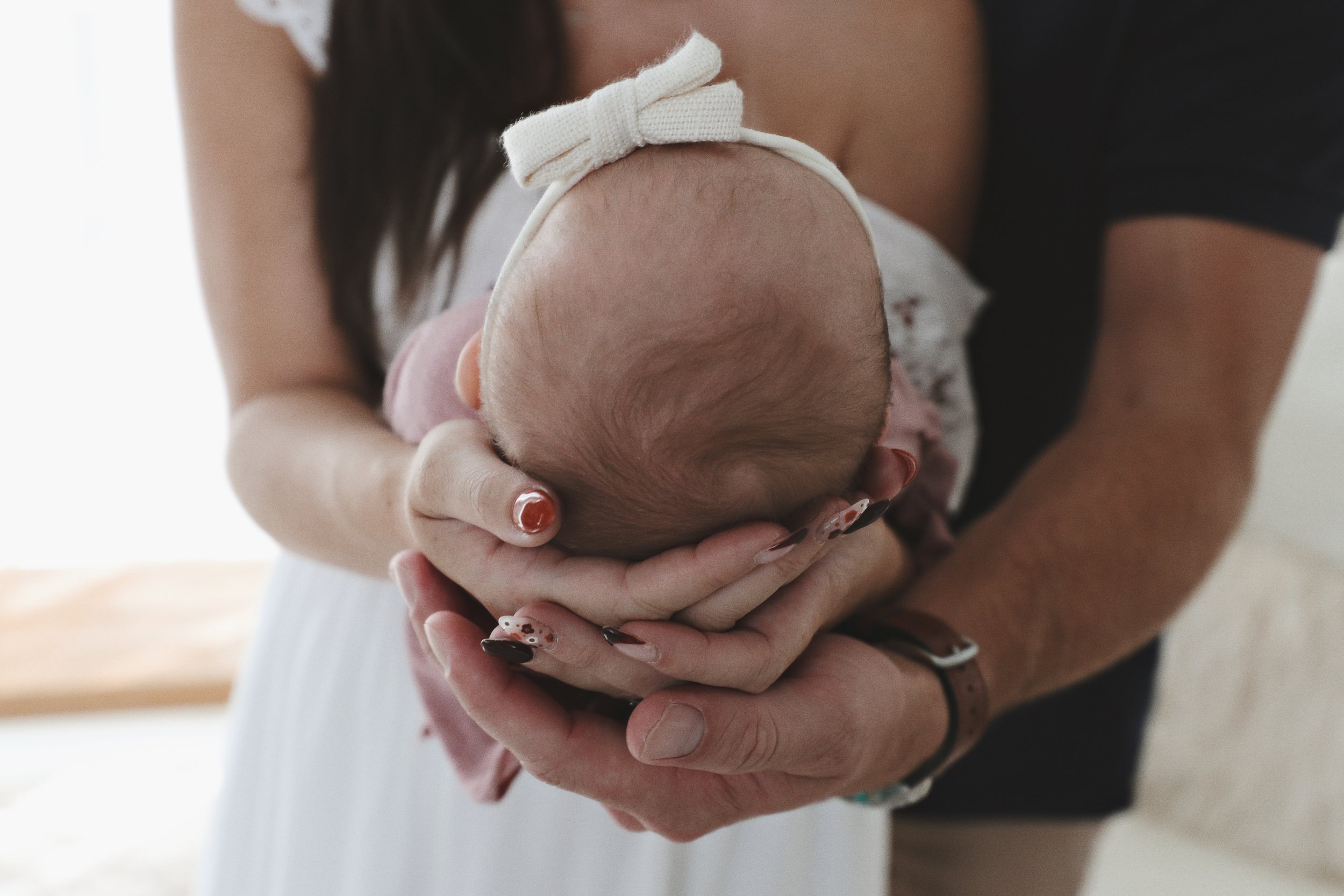 A woman holding a baby with her hands, supporting the baby's head and body. The baby is wearing a small bow on the head and is dressed in a pink outfit. The focus is on the baby's head and the woman's hands, with the woman wearing a dark top and a br