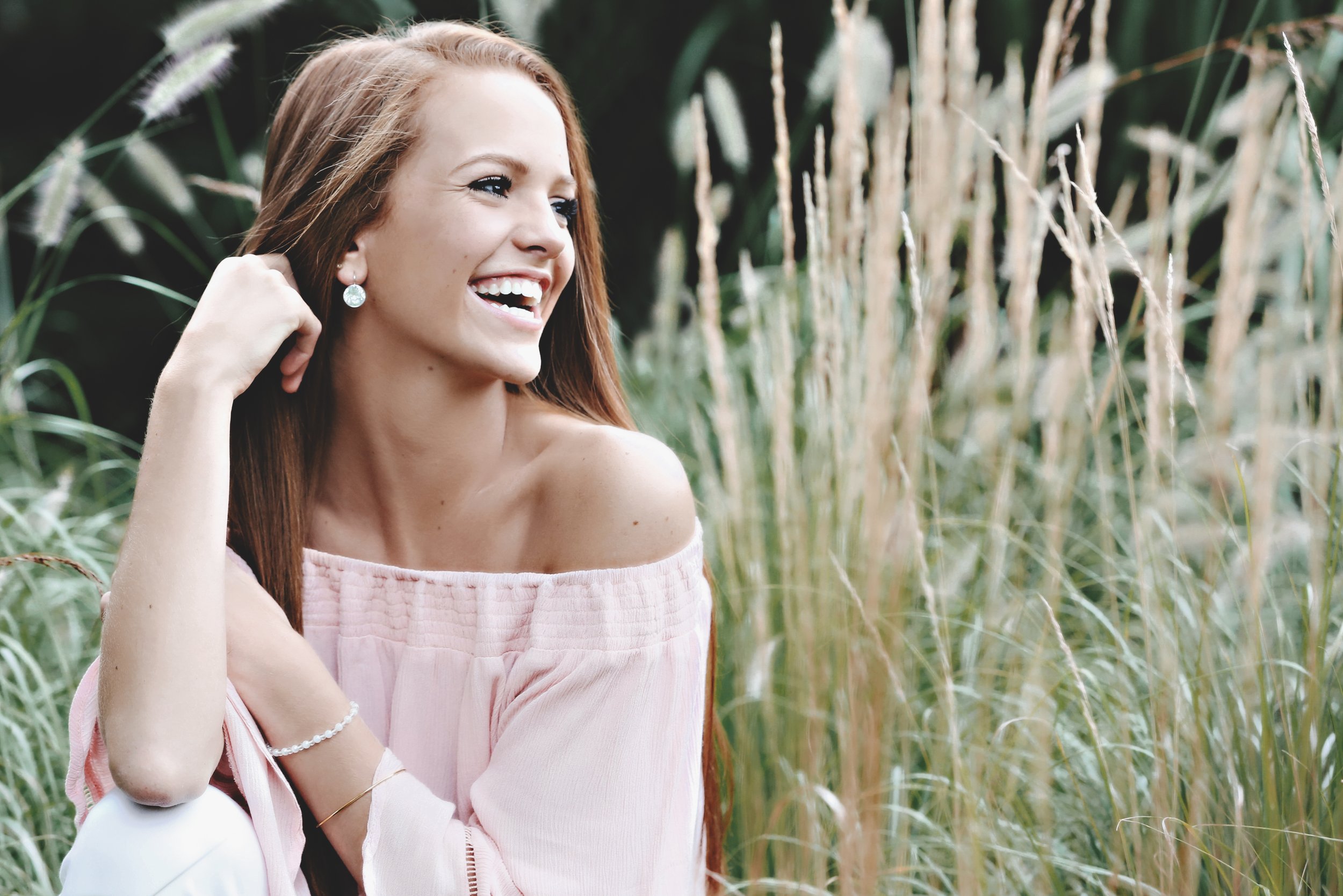 A woman with long red hair, wearing a pink off-shoulder top and jewelry, smiling and sitting outdoors among tall grass and greenery.