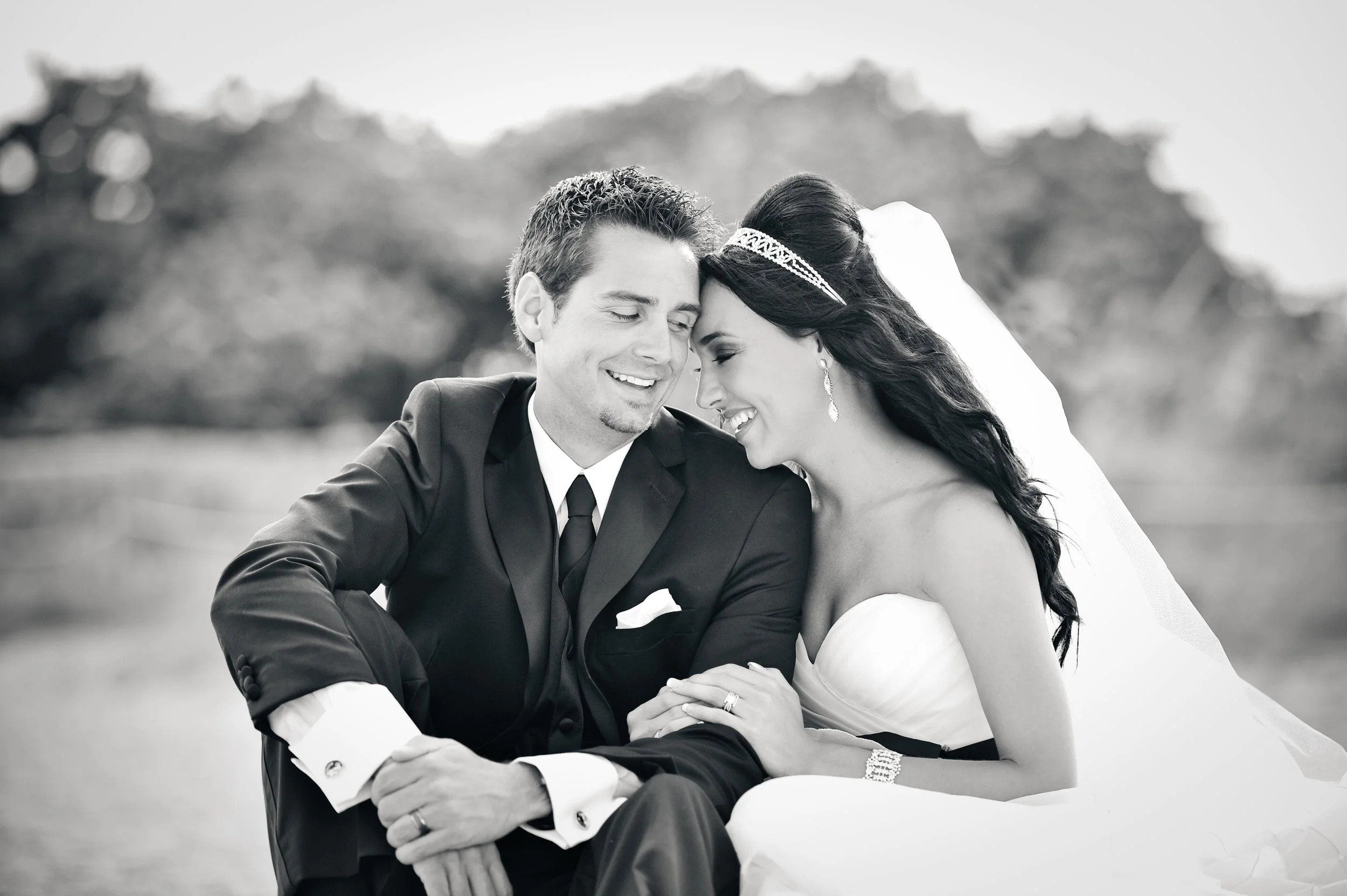 Black and white photo of a bride and groom sitting close together outdoors, smiling and touching foreheads, dressed in wedding attire.