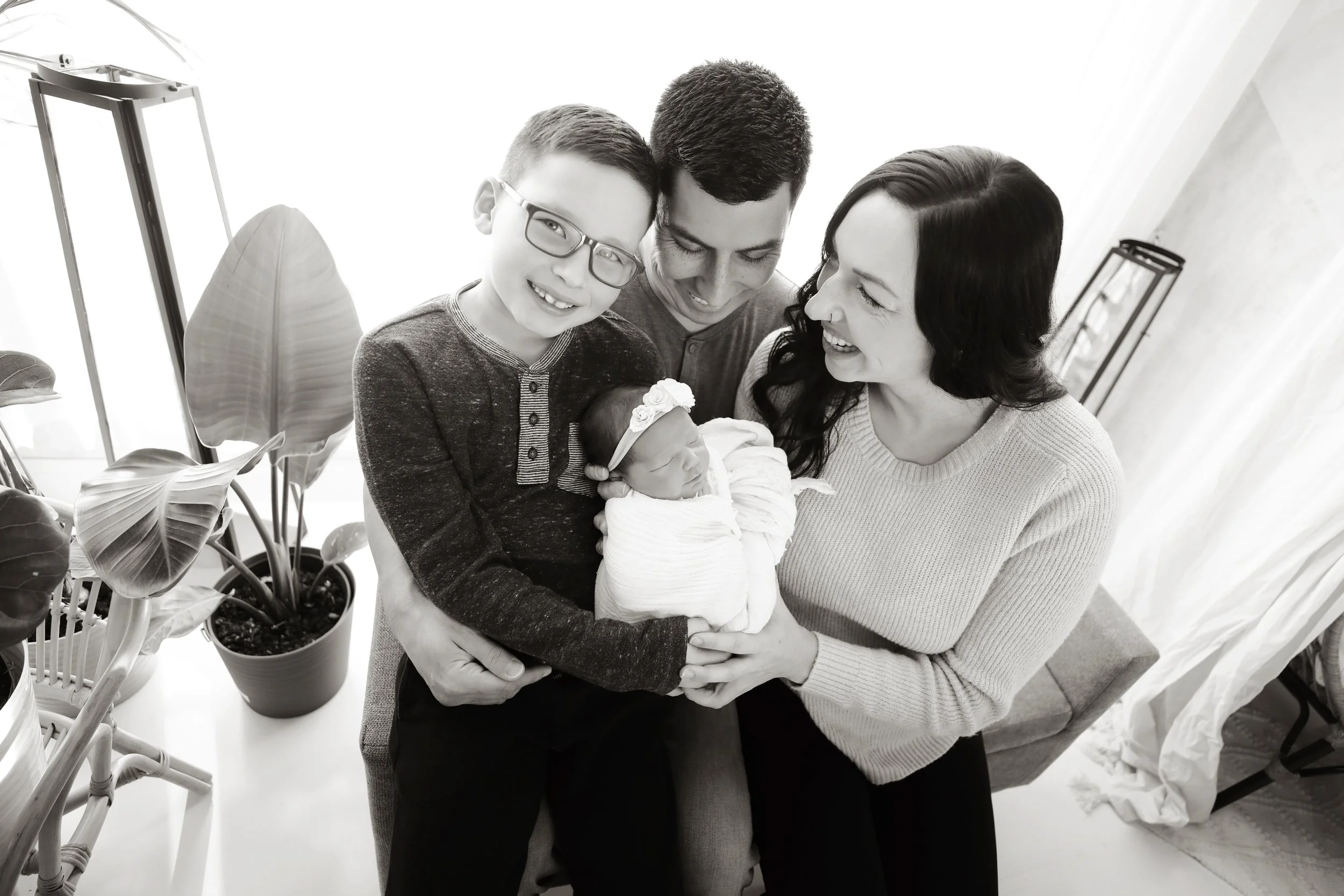 A family of four holding their newborn baby, smiling and looking at each other in a well-lit room.